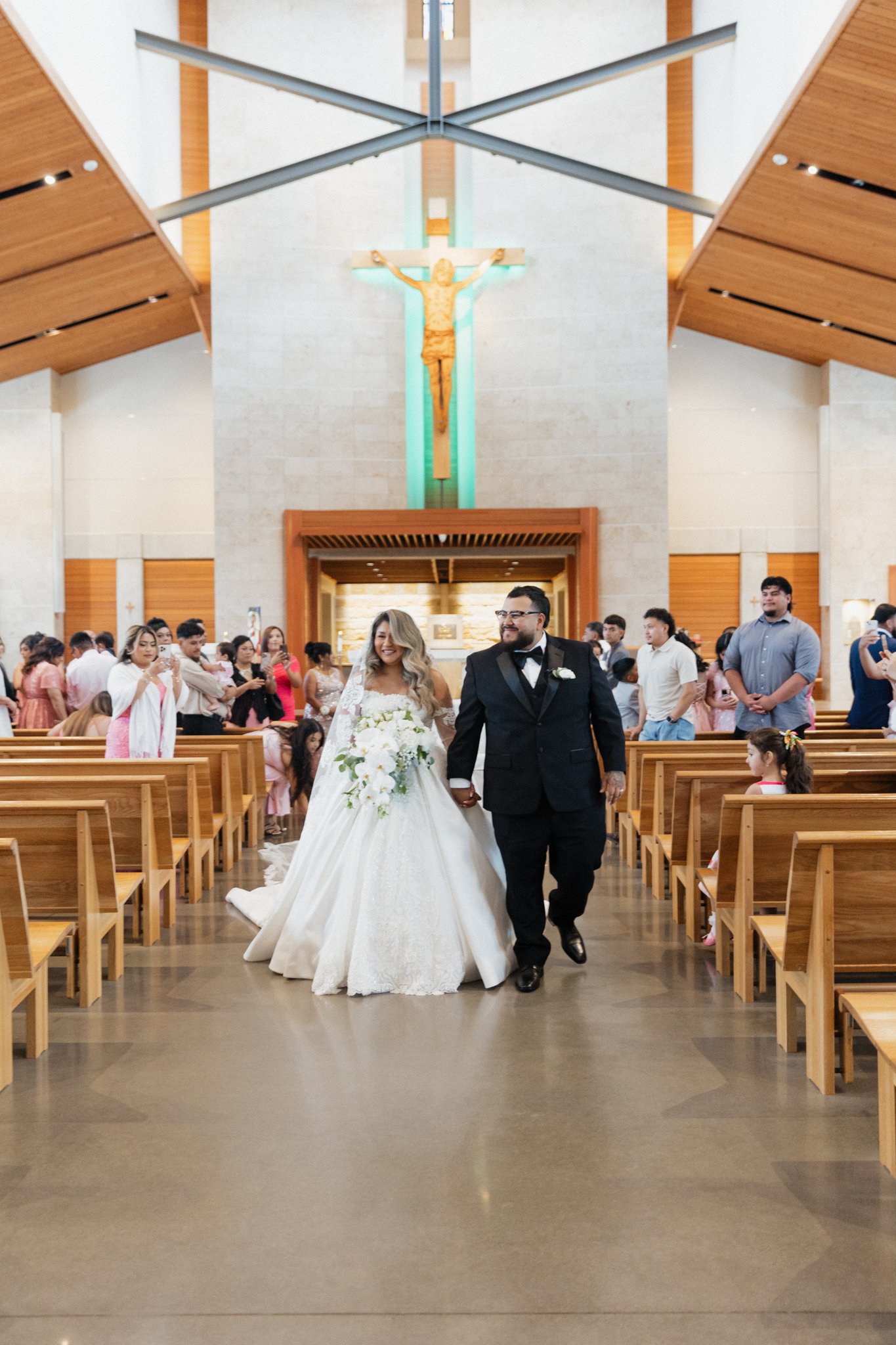 A bride and groom walking down the aisle of a church after their wedding ceremony, with guests watching and taking photos in the background, and a large crucifix hanging on the wall behind them.