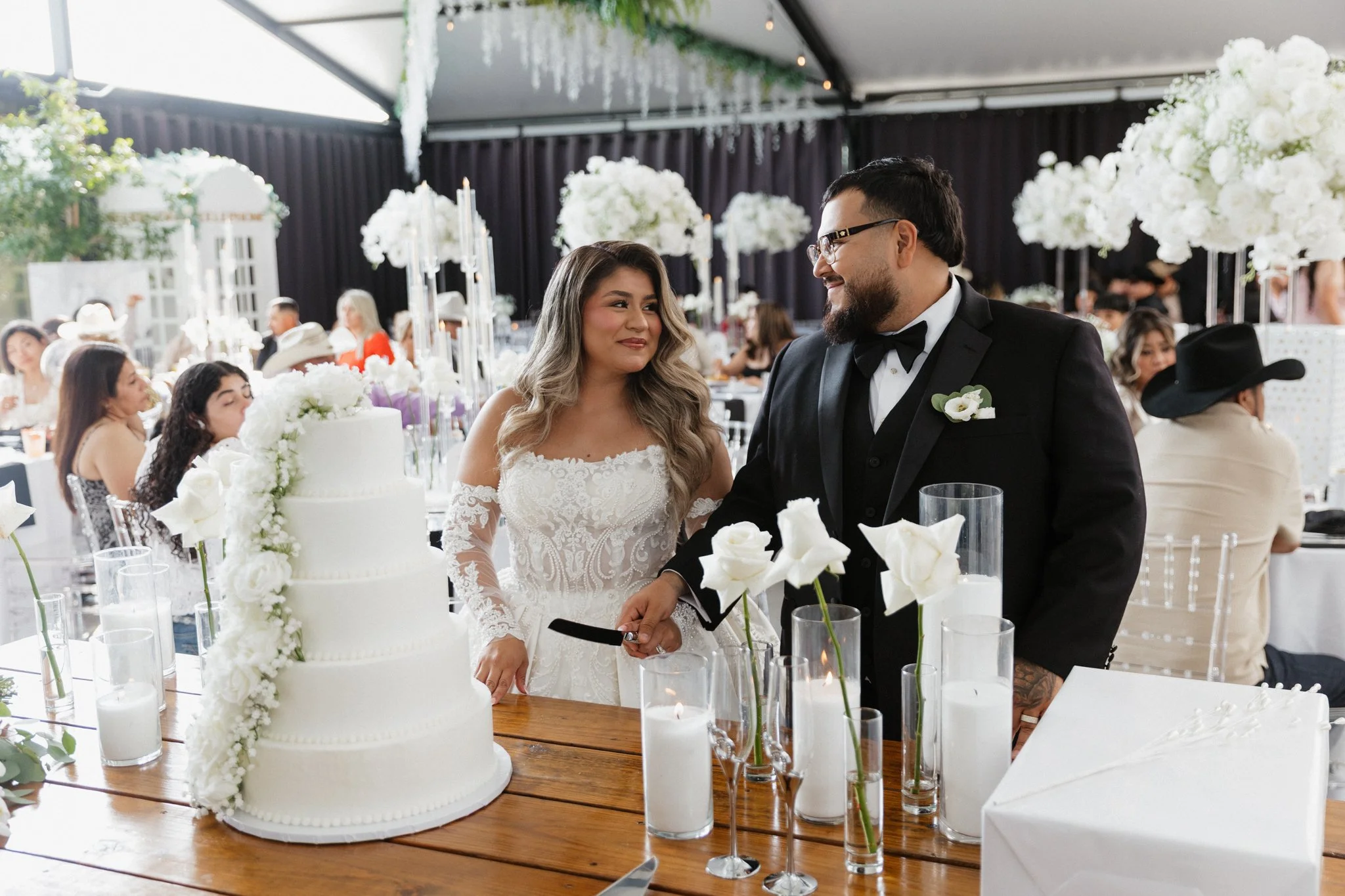 A bride and groom at their wedding reception, standing near a large white wedding cake decorated with white flowers. The bride is in a lace wedding dress, and the groom is in a black tuxedo with glasses. There are guests seated at tables with white a