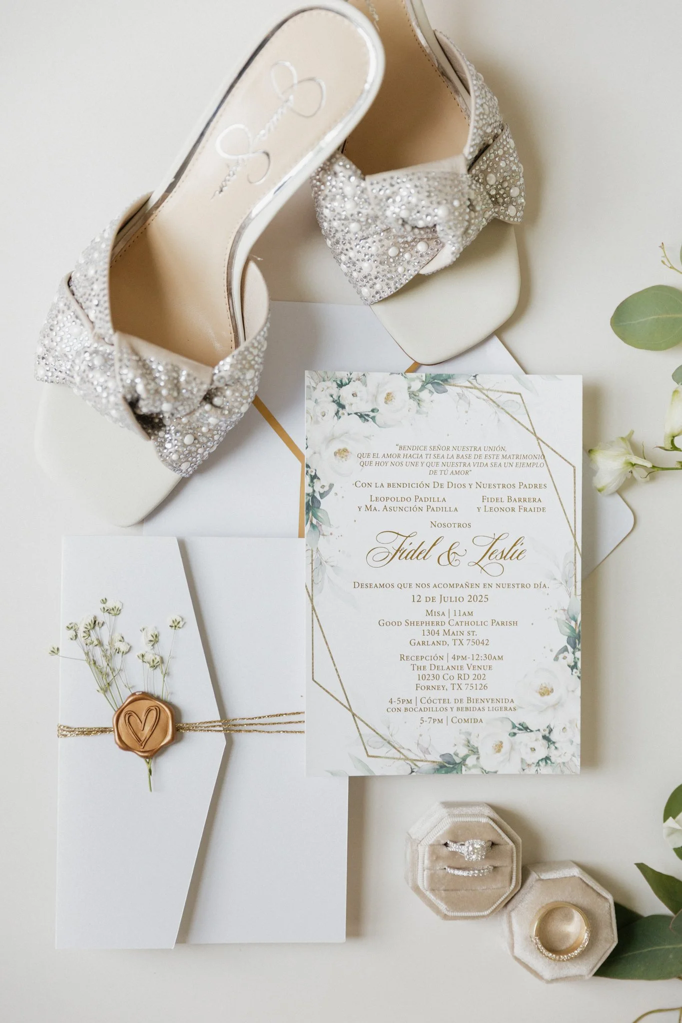 A pair of white high-heeled shoes with pearl and rhinestone embellishments on the bows, an elegant wedding invitation with floral design and gold accents, an envelope sealed with a wax stamp featuring a heart, and two rings in a jewelry box, all arra