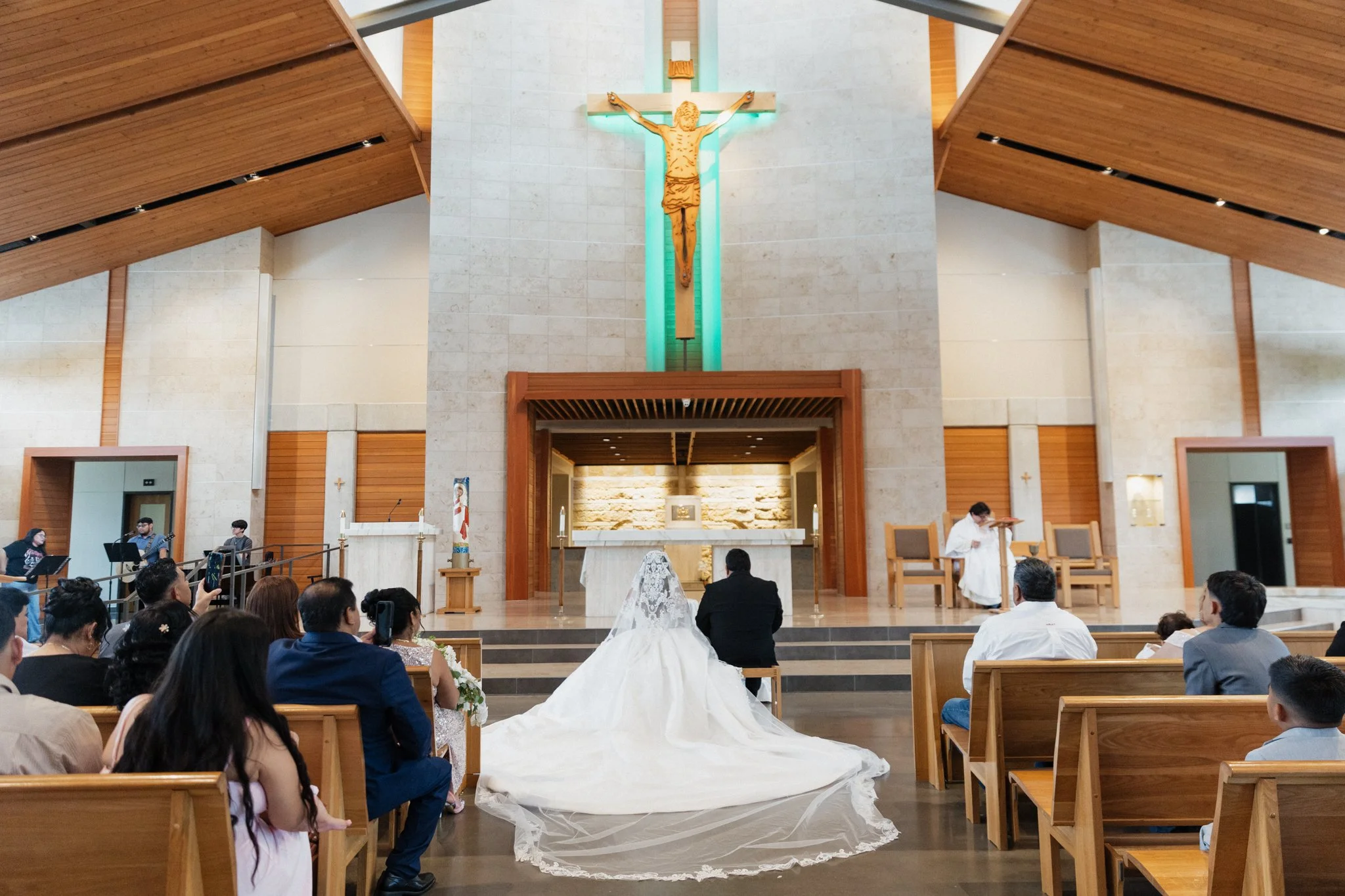 A wedding ceremony in a church with a bride in a white gown and veil, and a groom in a black suit, seated in front of the altar facing the minister. The church has wooden accents and a large crucifix on the wall behind the altar. Guests are seated on