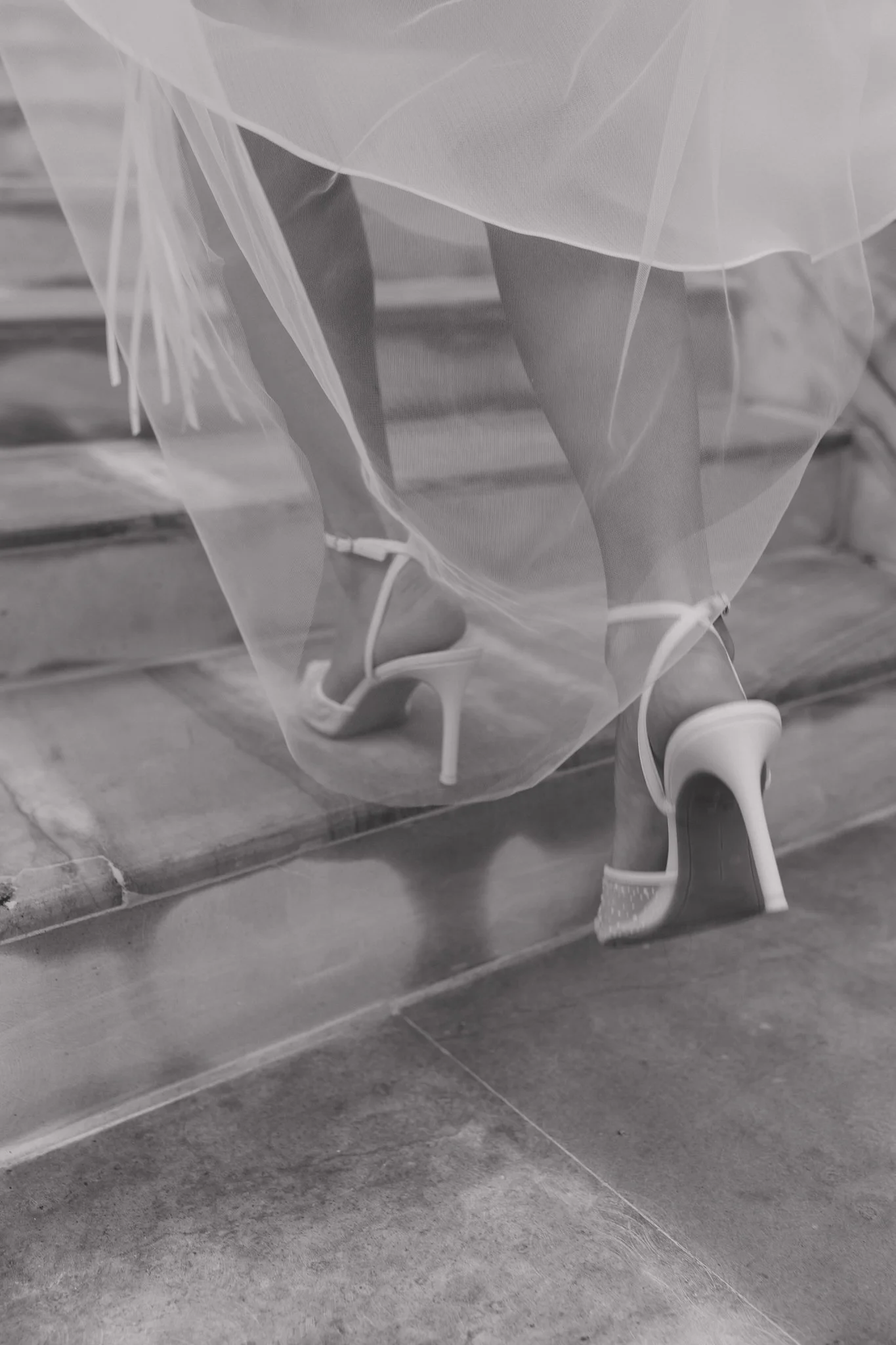 Close-up of a woman's legs and feet wearing high-heeled sandals and a flowing dress, seen through a sheer veil, walking on a tiled staircase.