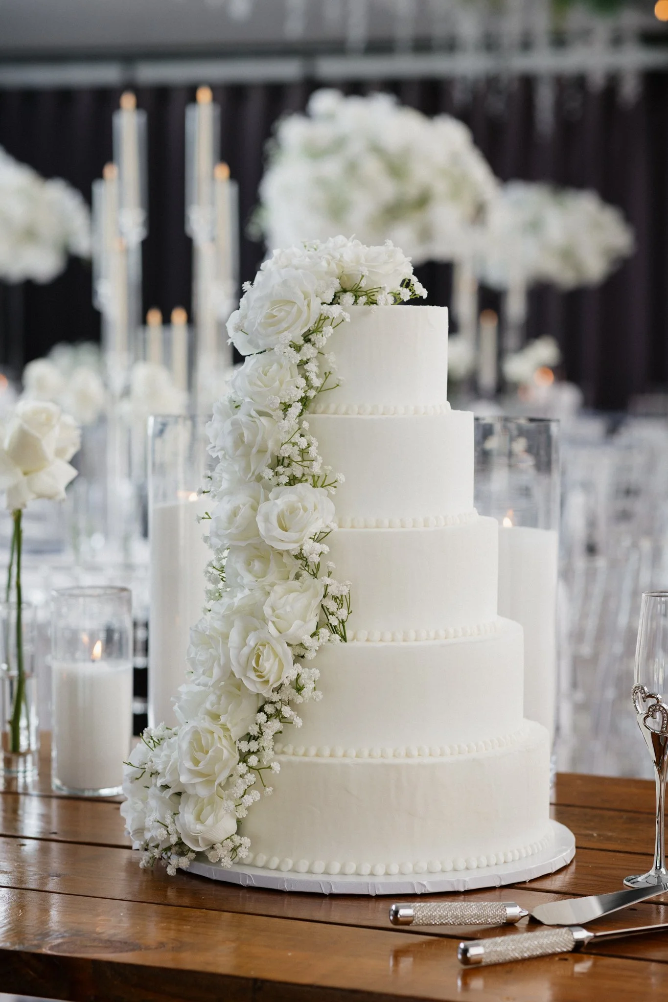 A five-tiered white wedding cake decorated with white flowers, placed on a wooden table at a wedding reception.