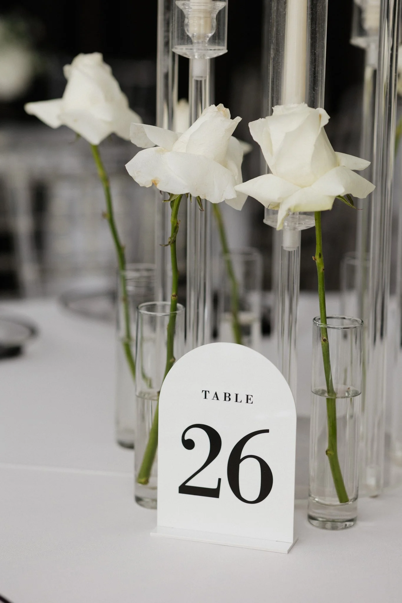 White roses in glass test tubes with a table number card reading 'Table 26' on a white tablecloth.