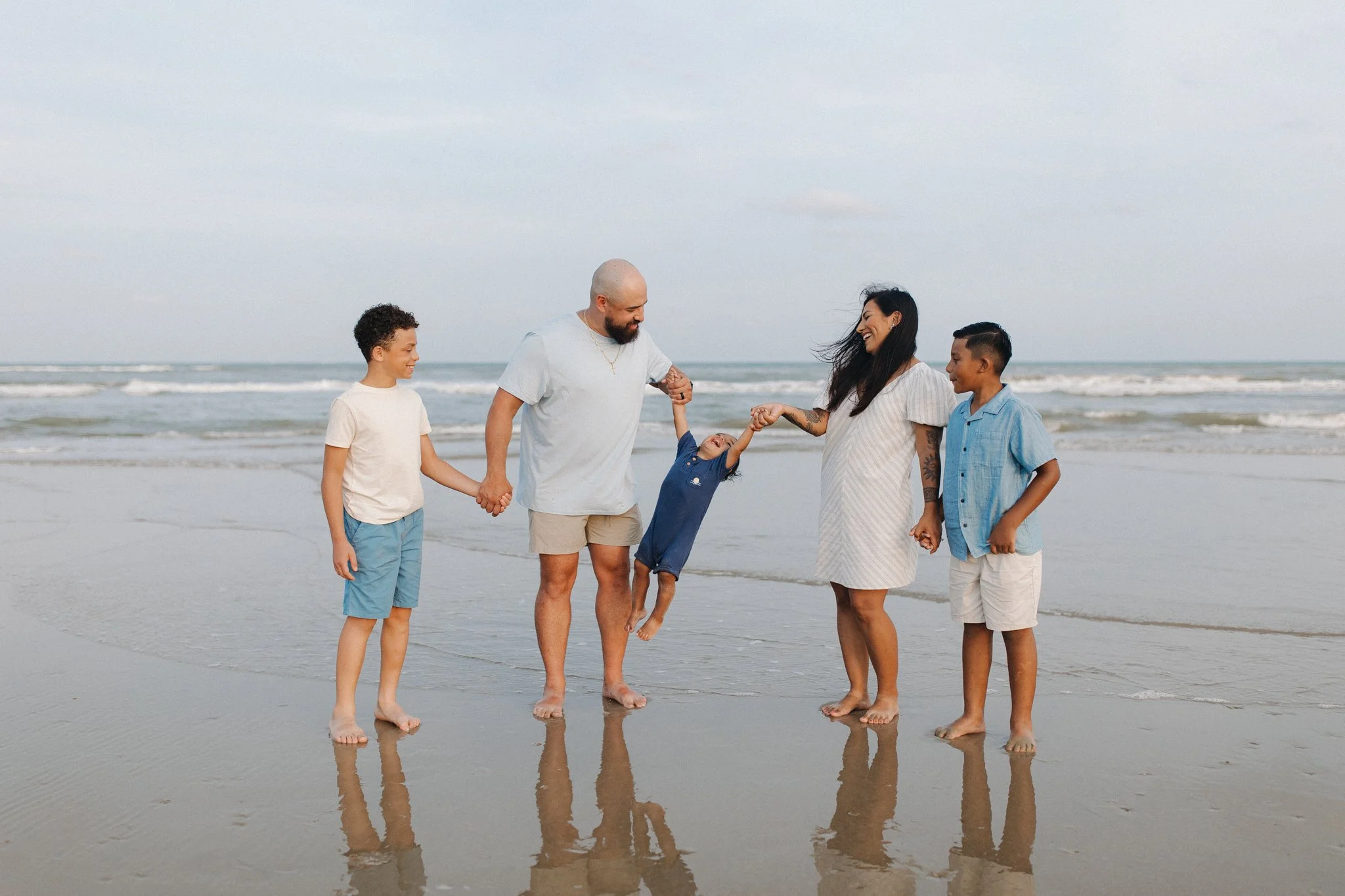 A family of six, including two adults and four children, holding hands and enjoying a walk on the beach with ocean waves in the background. They are smiling and dressed in casual summer clothing.