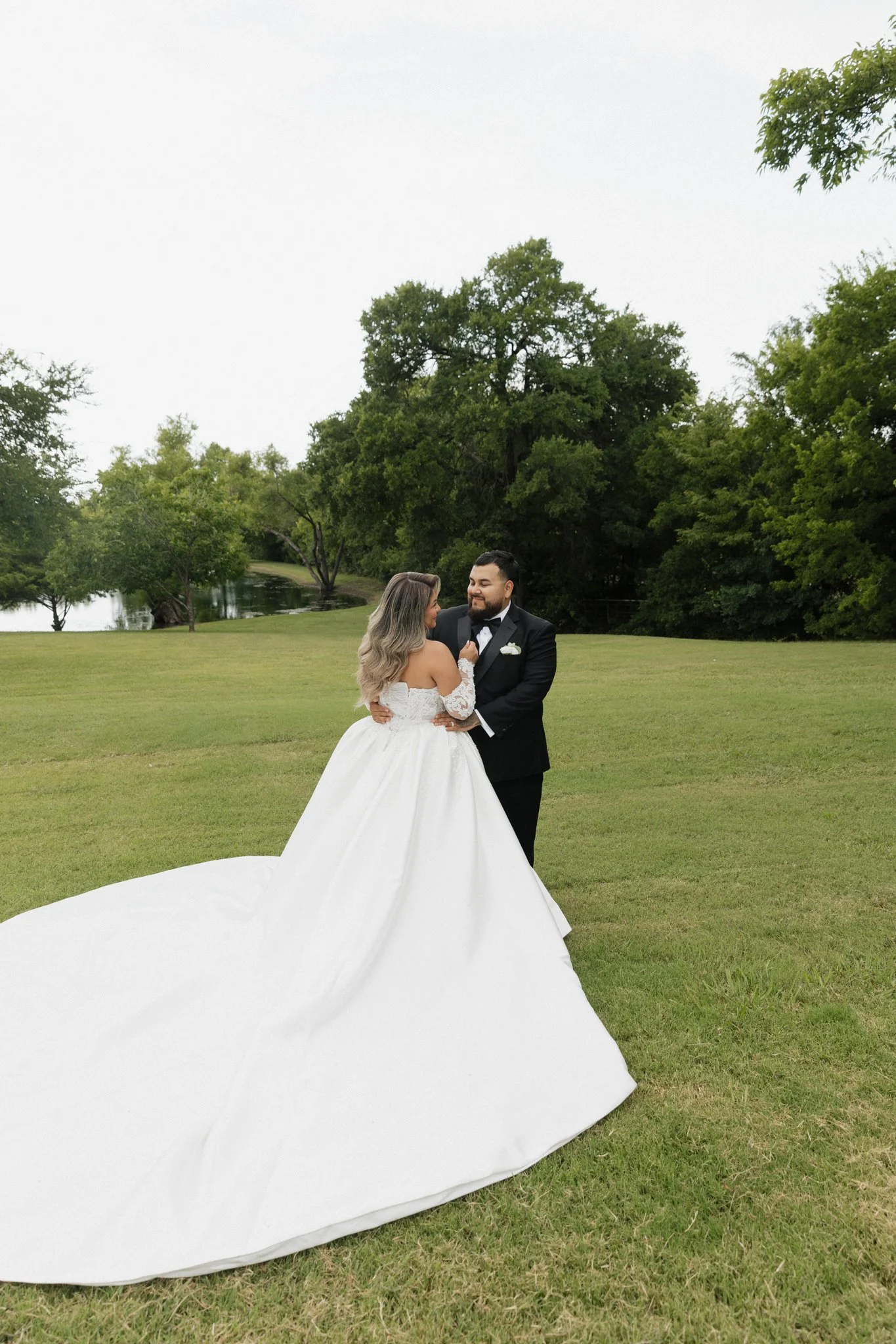 Bride and groom dancing outdoors on grass, with trees and a pond in the background, during a wedding celebration.