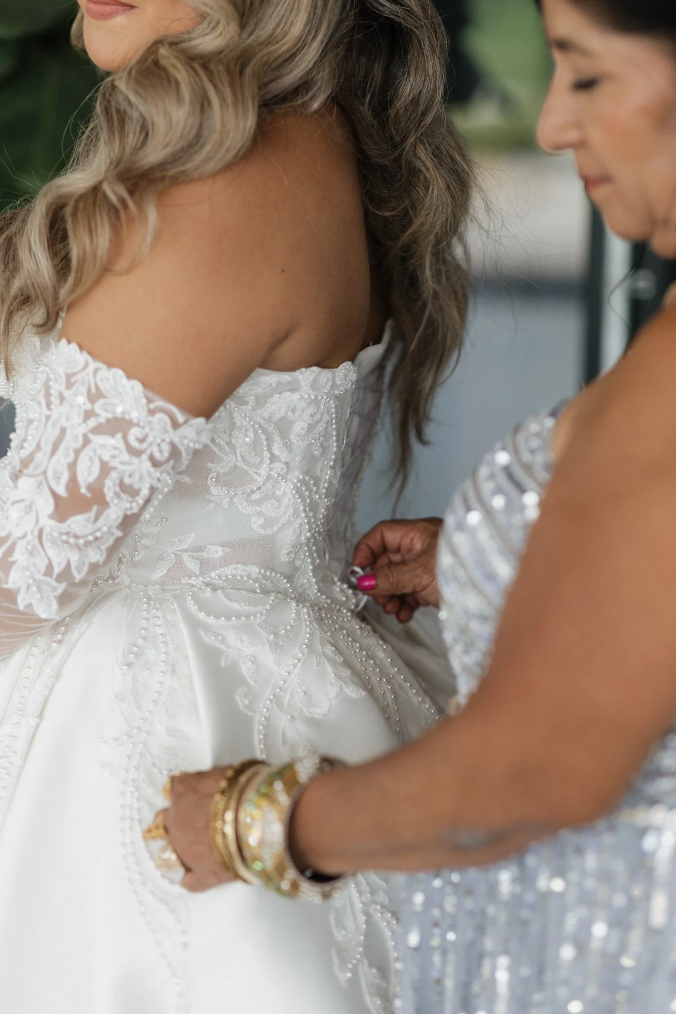 A woman in a strapless, lace and embroidered wedding dress is being helped with her dress, possibly on her wedding day, by another woman dressed in a silver, sequin-covered outfit.
