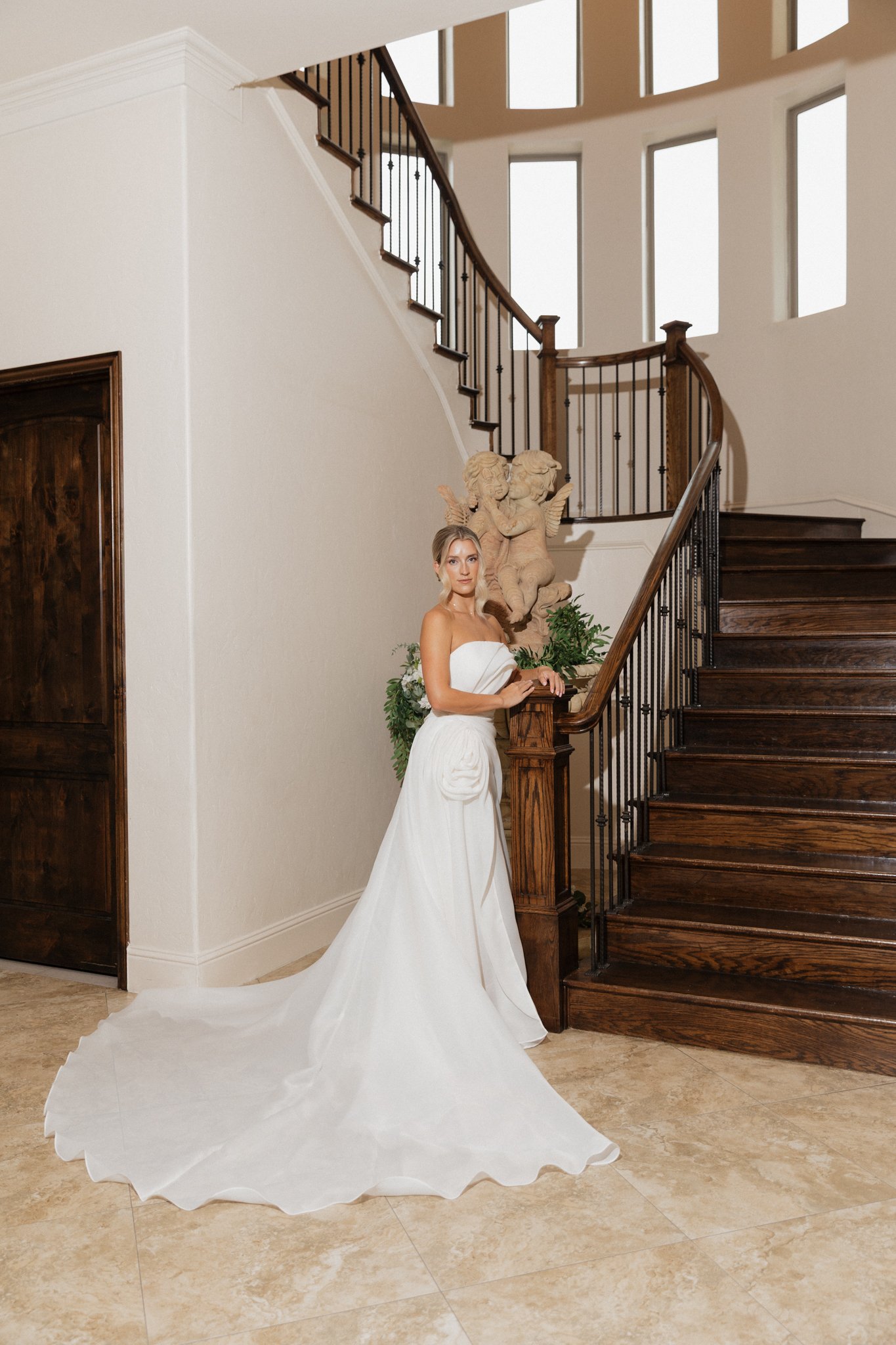 A woman in a white wedding dress stands at the bottom of a wooden staircase in a well-lit indoor setting, near a decorative stone cherub sculpture and green plants.