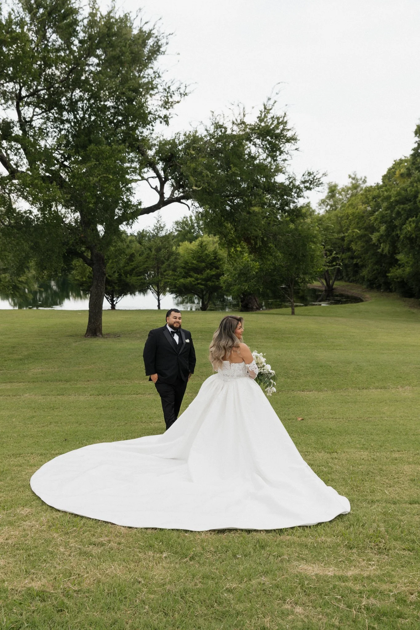 A bride in a white wedding gown holding a bouquet walking across a grassy field while a groom in a black tuxedo watches her. Trees and a lake are in the background.