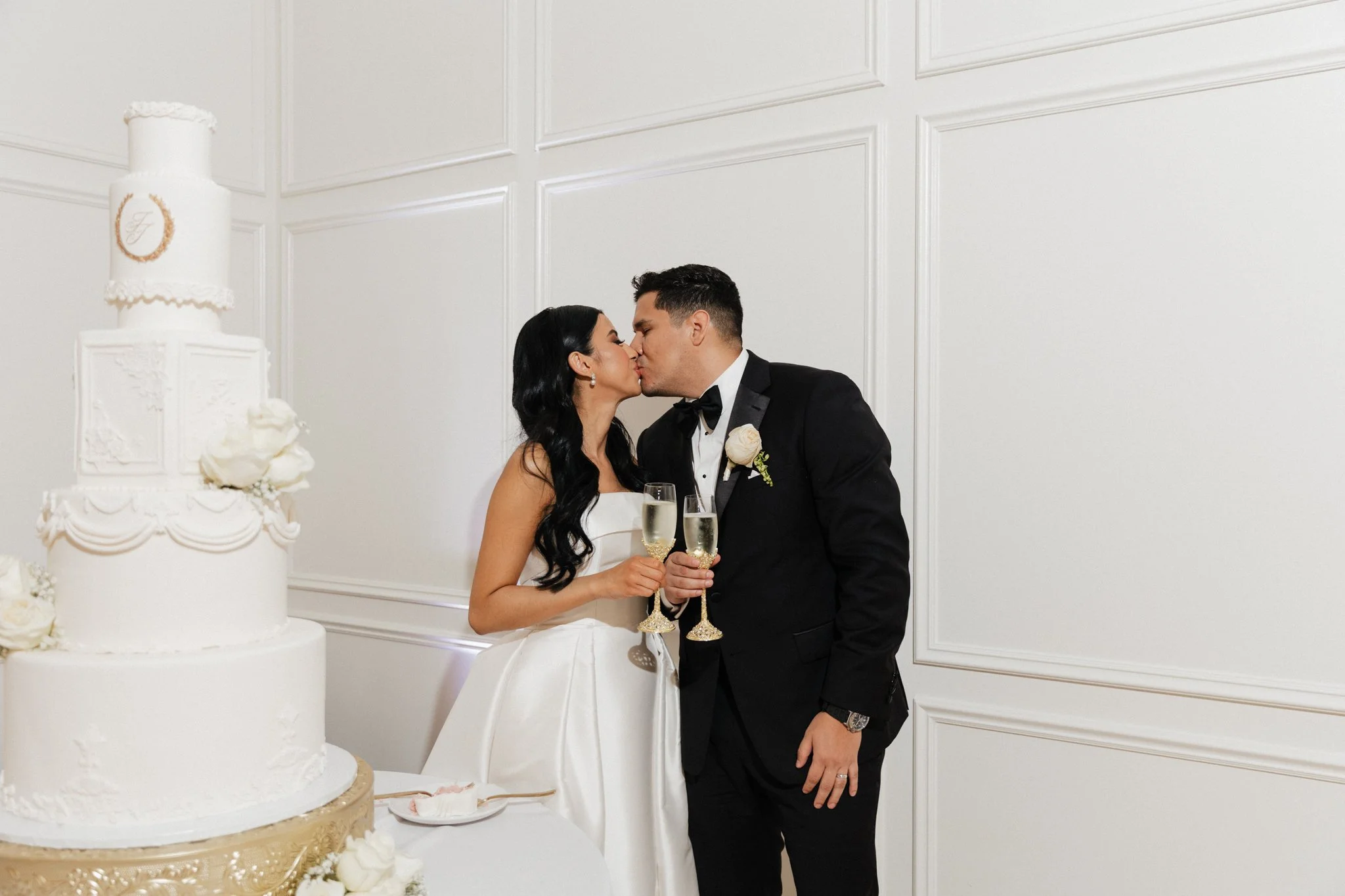 A bride and groom share a kiss at their wedding reception, holding champagne glasses, with a tall white wedding cake decorated with white flowers in the foreground.