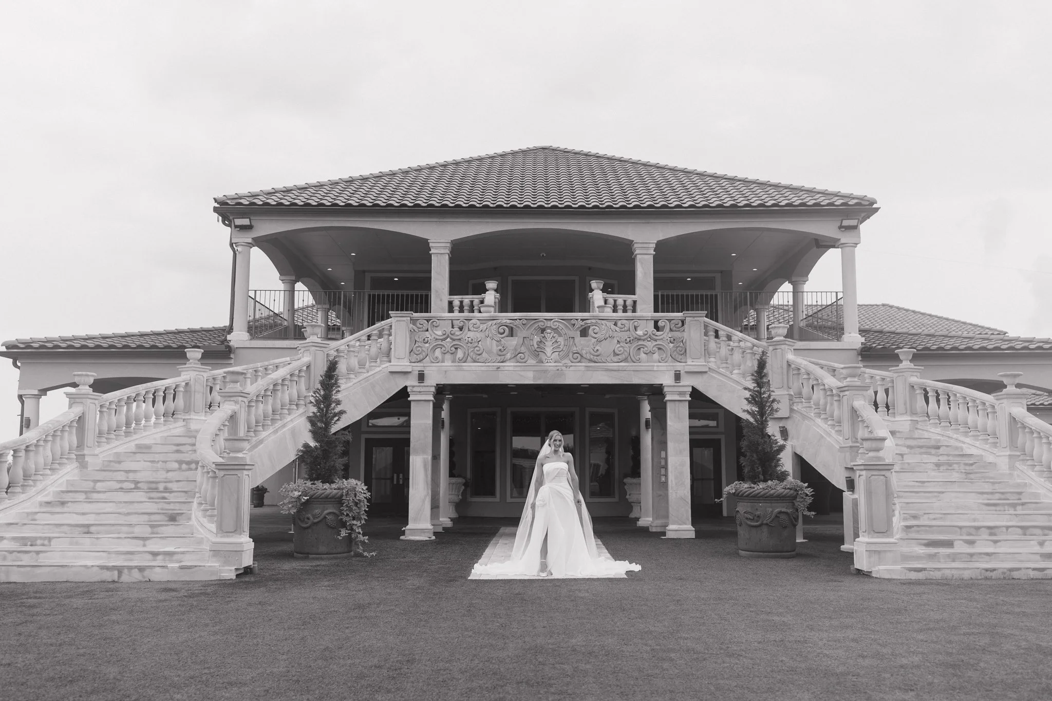 A bride in a wedding dress standing in front of a large, elegant house with stairs and a balcony, in black and white.