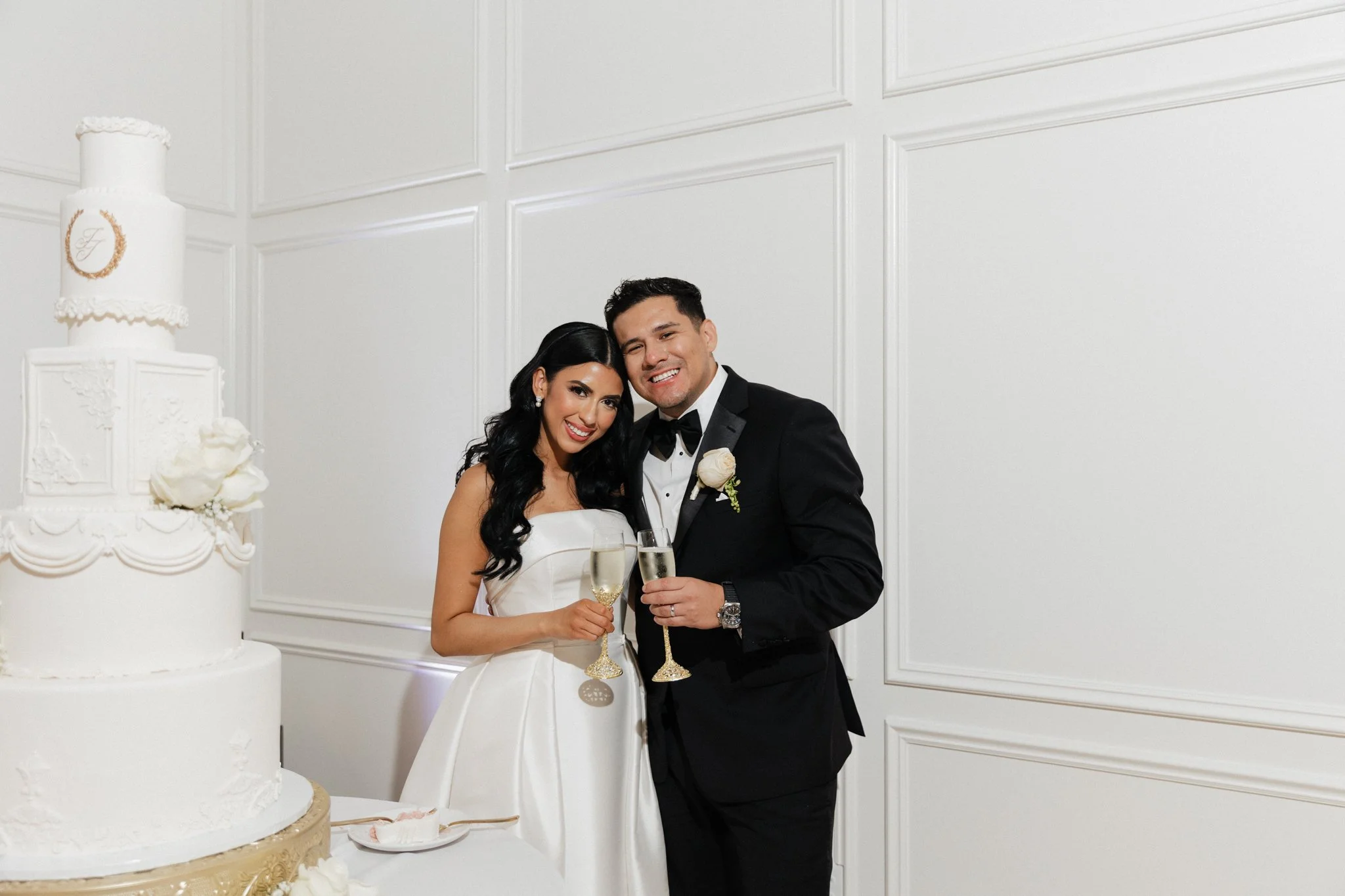 A newlywed couple in wedding attire, celebrating with champagne, standing close together in front of a white wall, with a tiered wedding cake decorated with white flowers and intricate designs seen on the left side.