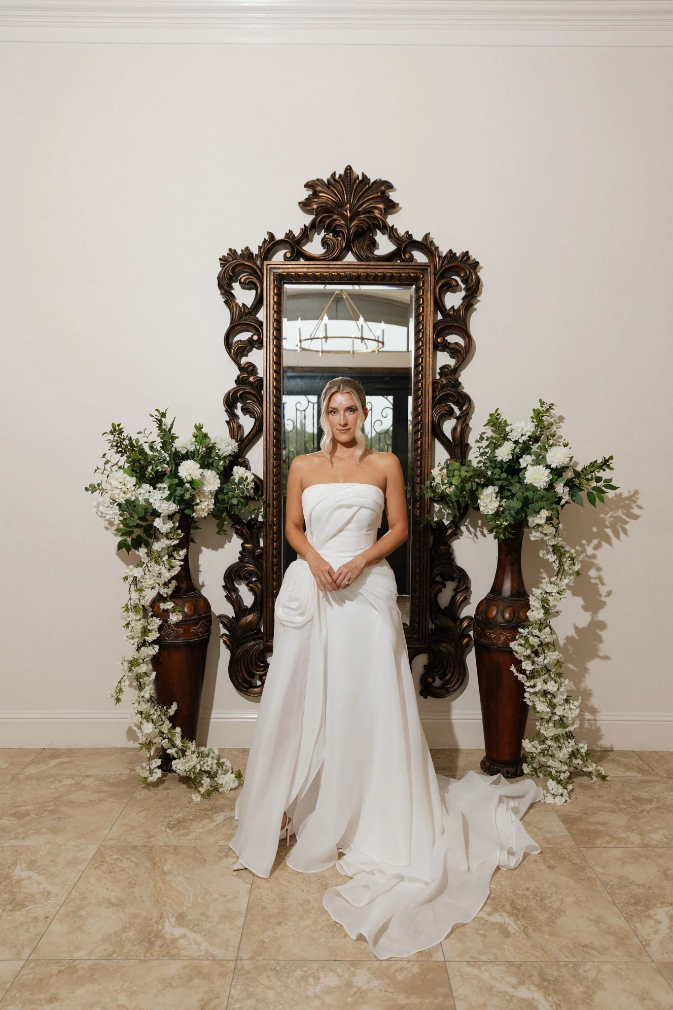 A bride in a white wedding dress standing in front of a large ornate mirror with flowers on each side.