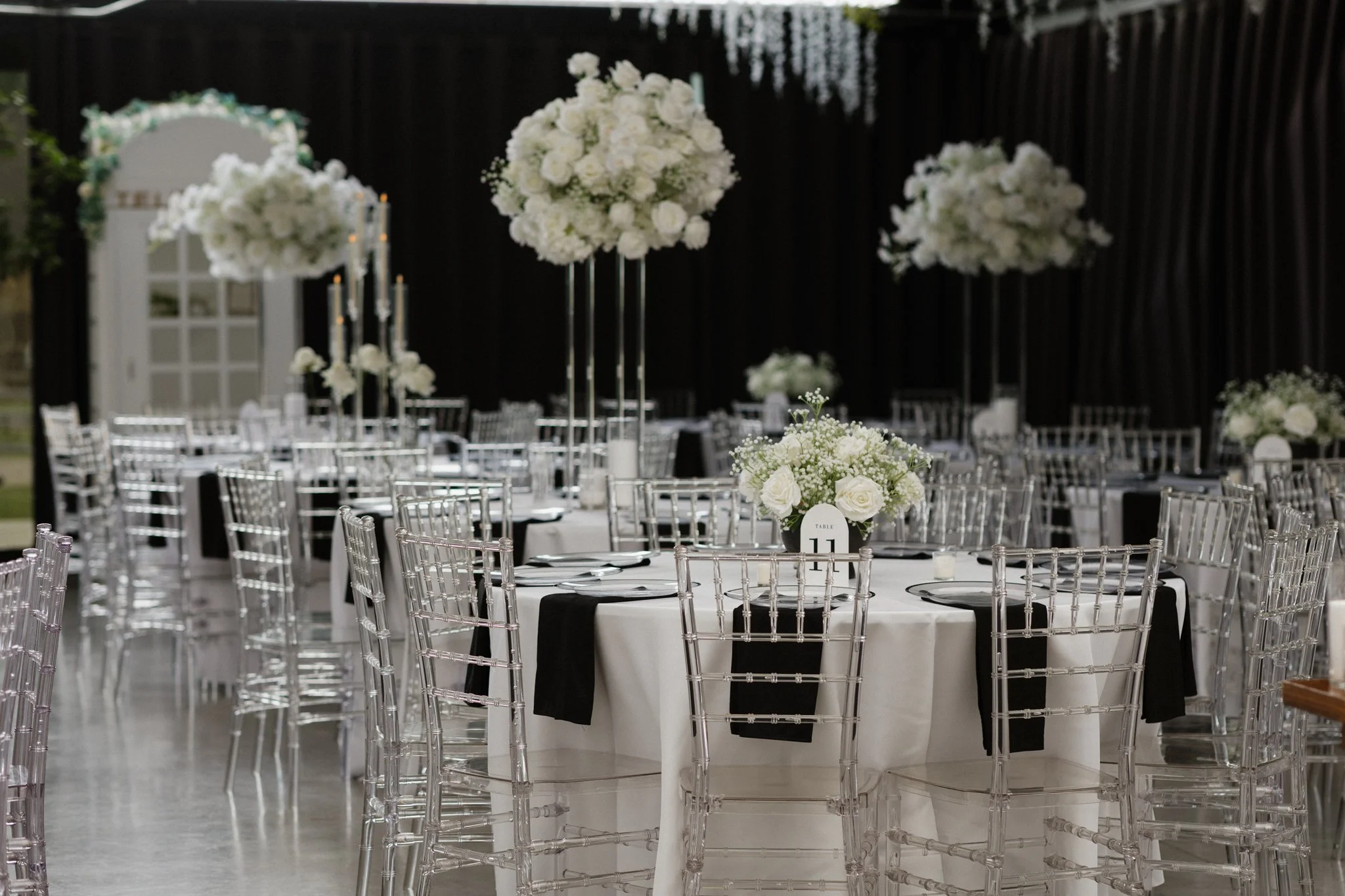 Elegant event reception with round tables, clear acrylic chairs, and floral centerpieces of white roses and baby's breath, set against a dark curtain backdrop.