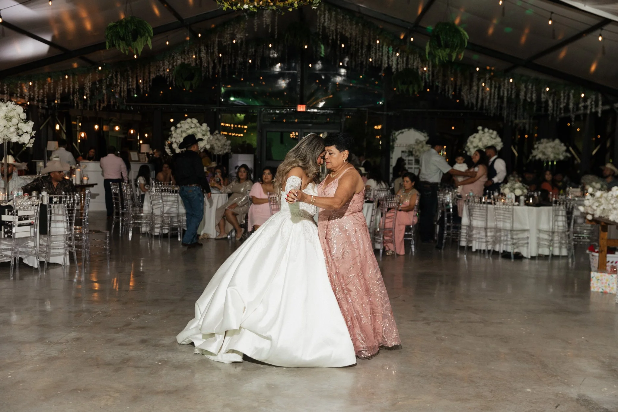 A bride in a white wedding gown dancing with an older woman in a pink dress at a wedding reception. The venue has floral decor, white tablecloths, and guests seated at tables.