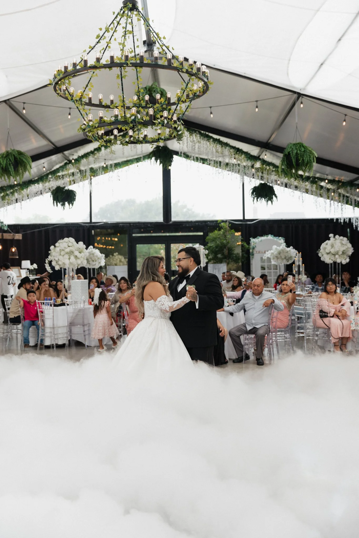 A bride and groom dancing at their wedding reception inside a decorated event tent, with guests seated at tables in the background and greenery hanging from the ceiling.