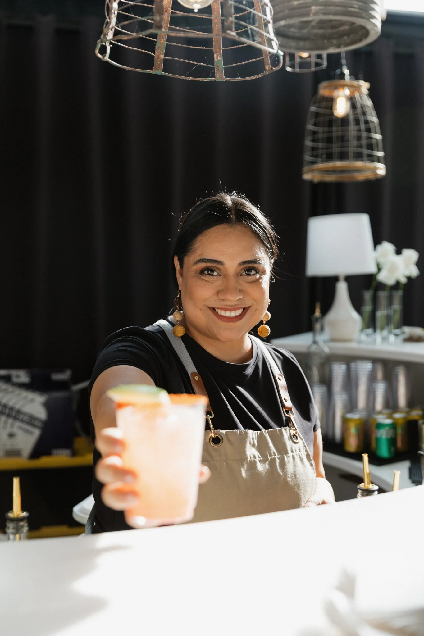 A woman with dark hair and earrings is smiling and holding a drink towards the camera. She is wearing a black shirt and a beige apron, standing behind a white bar counter with a dark background and decorative hanging baskets.