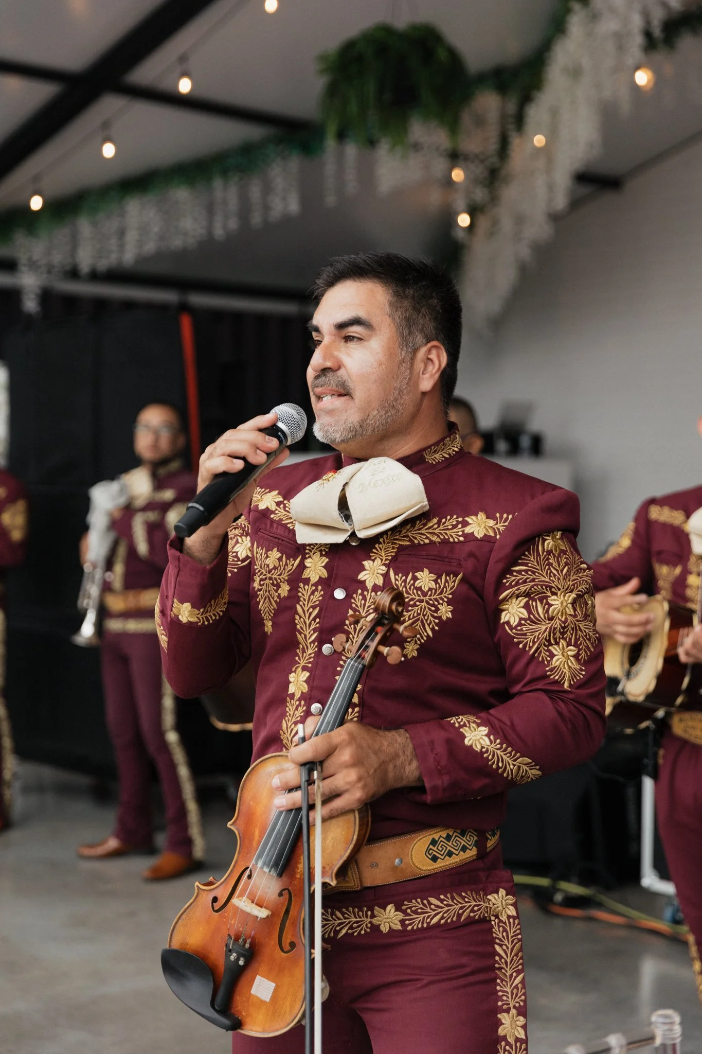 A man holding a microphone and a violin, wearing a maroon and gold embroidered traditional outfit, performing at an indoor event with background musicians.
