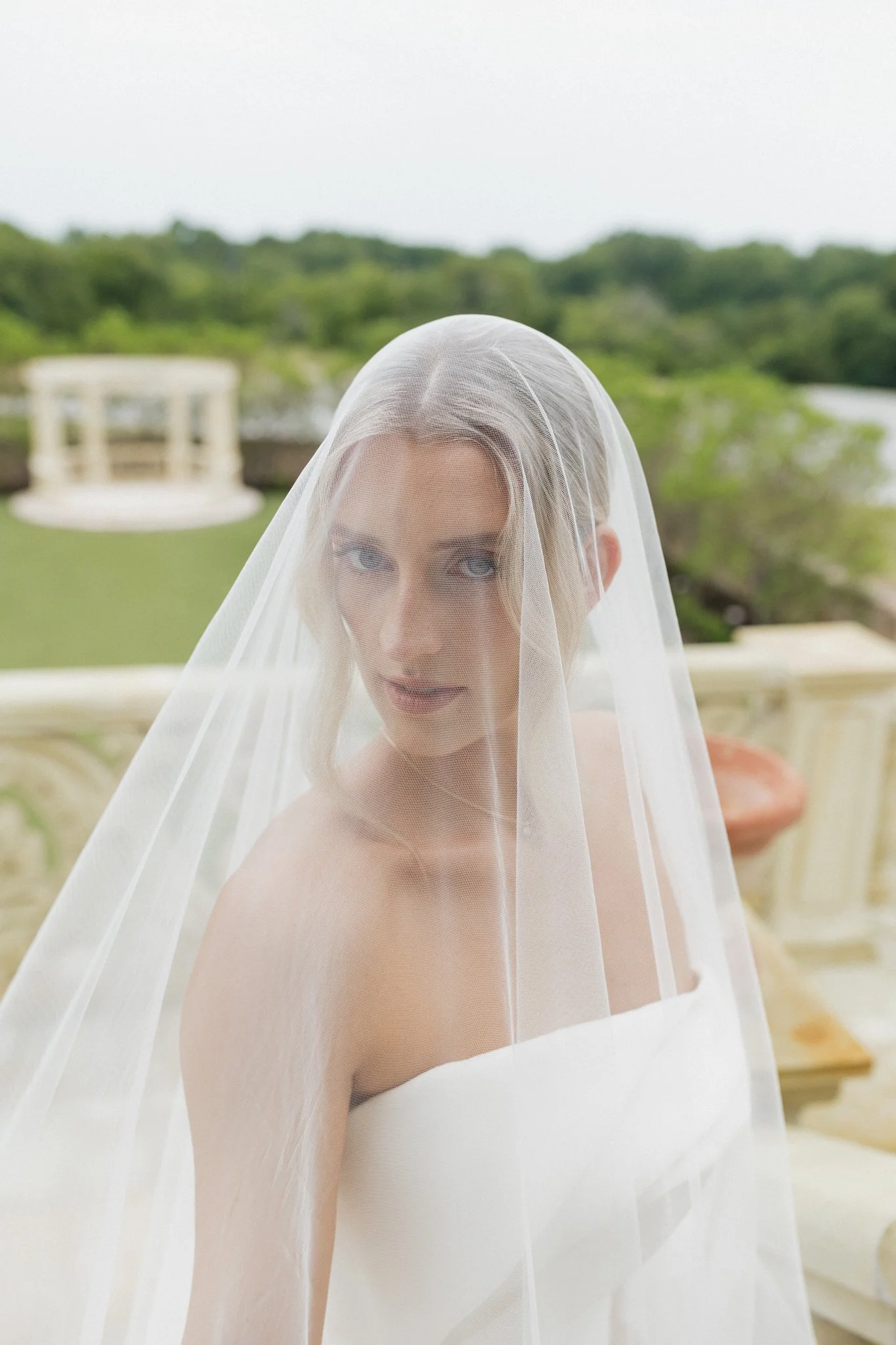 Woman with blonde hair wearing a white wedding dress and veil, standing outdoors with a garden and a gazebo in the background.
