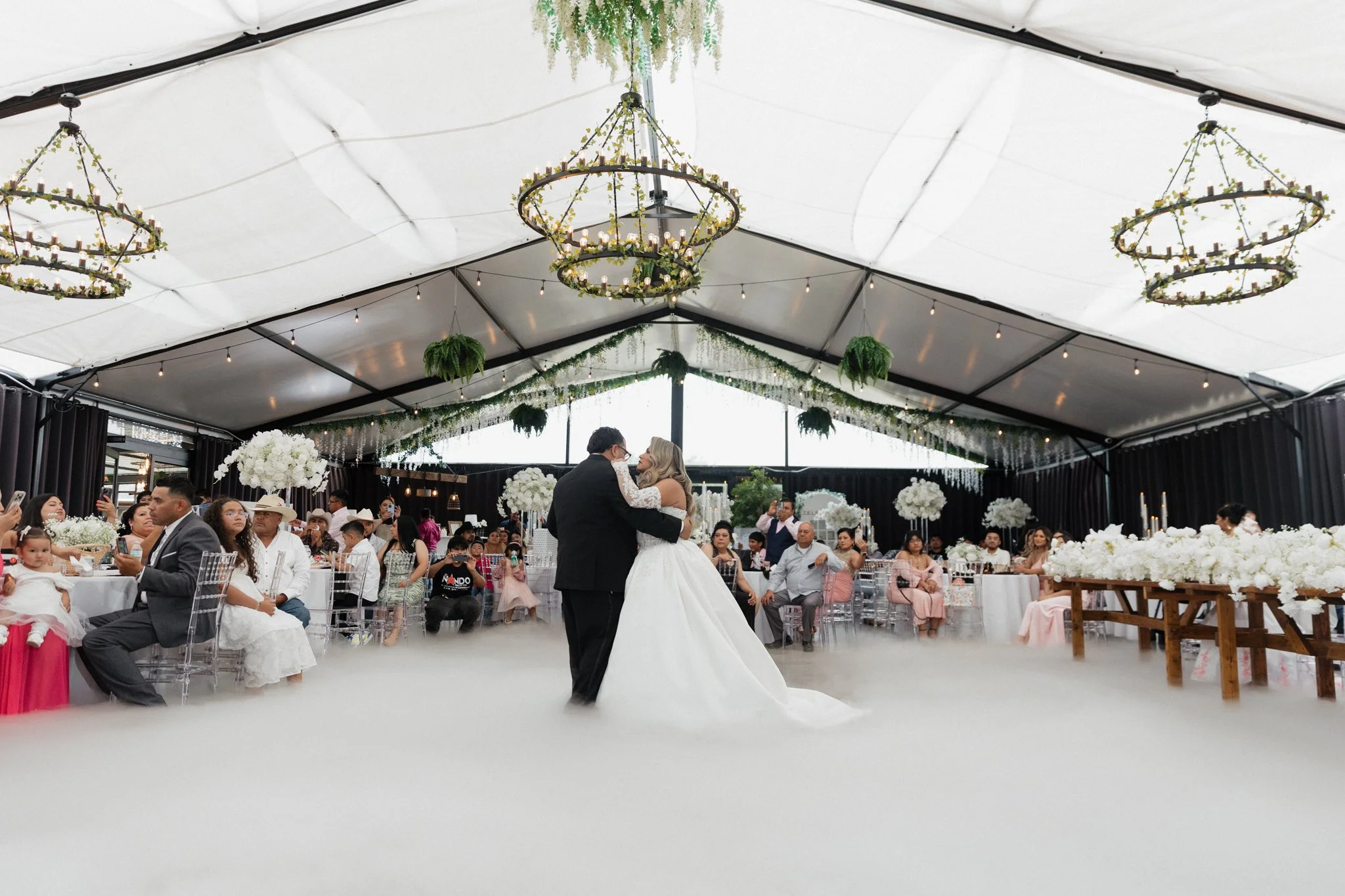Bride and groom dancing at their wedding reception under a decorated tent with hanging chandeliers and floral arrangements
