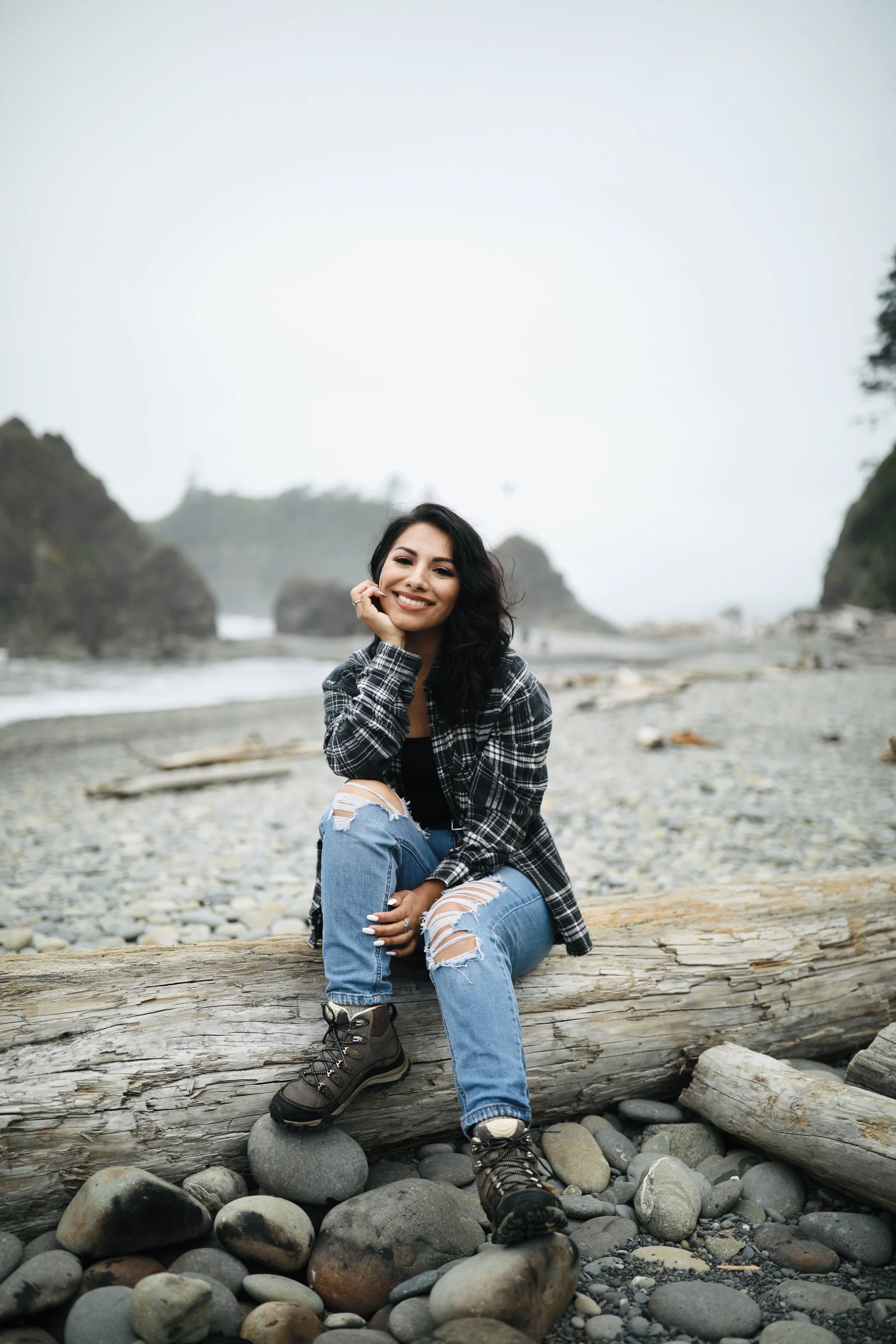 Young woman sitting on a log on a rocky beach with ocean waves, distant cliffs, and cloudy sky in the background.