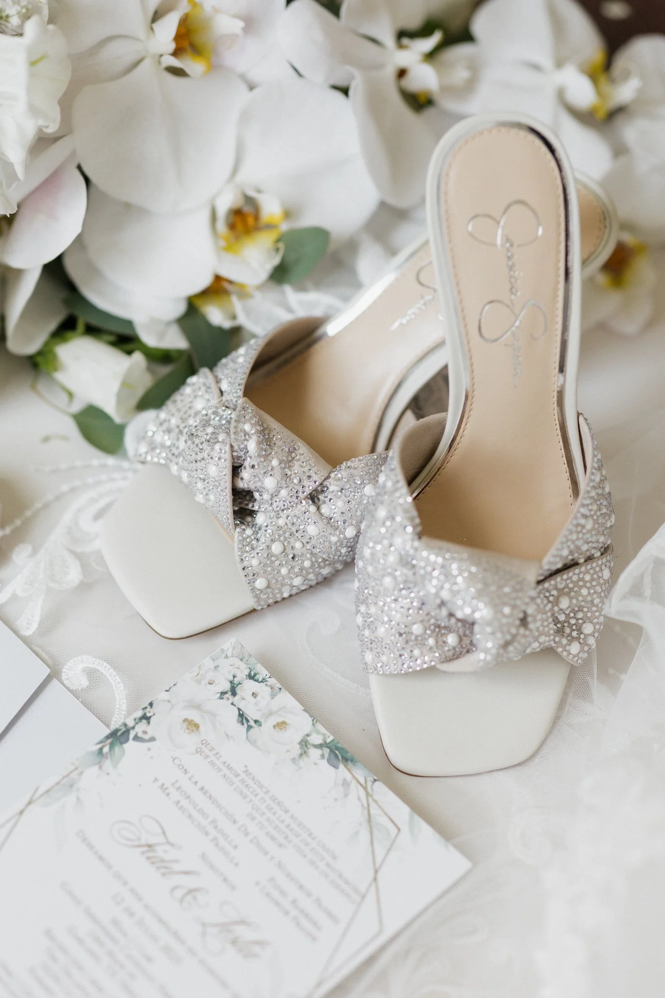 A pair of elegant, high-heeled wedding shoes adorned with rhinestones and pearls, placed on a white fabric with floral accents and a wedding invitation nearby.