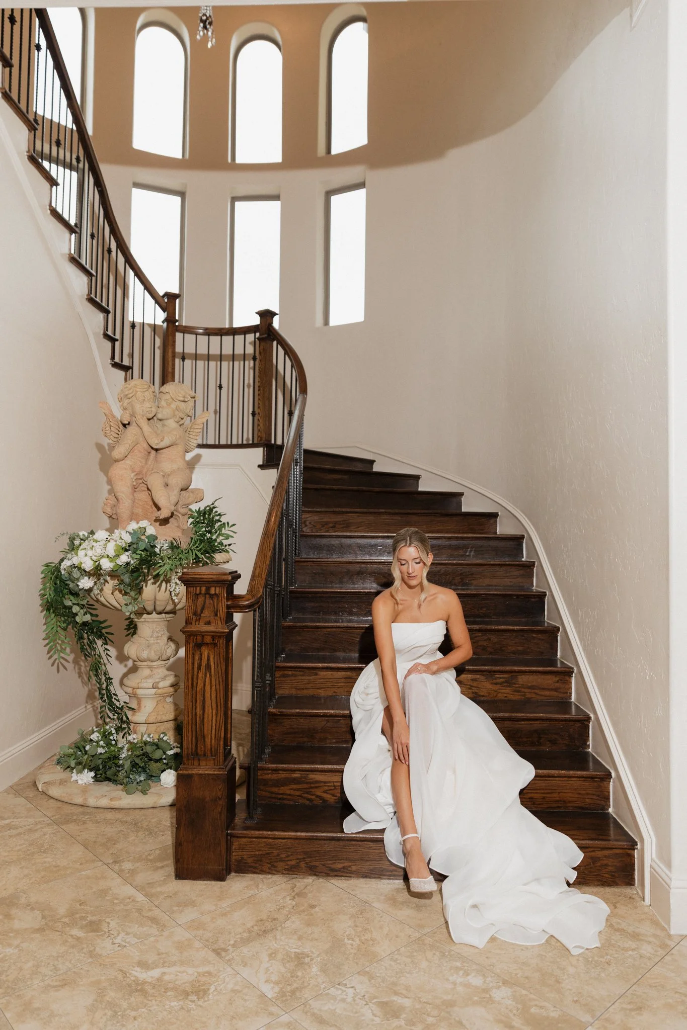 A young woman in a white wedding dress sitting on dark wooden stairs in a spacious, well-lit interior with high arched windows, next to a decorative stone pedestal with floral arrangements and a cherub sculpture.
