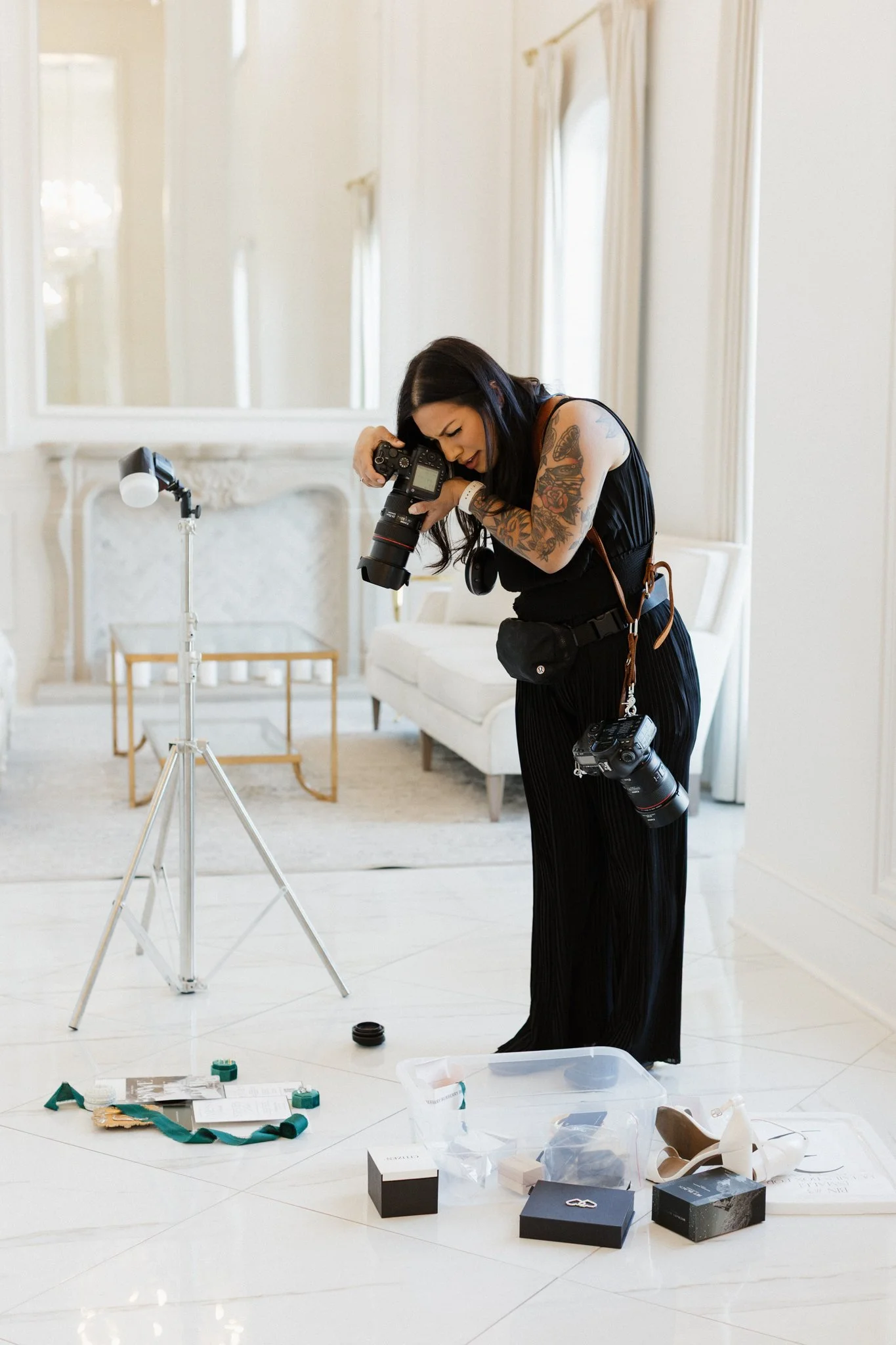 A woman with tattoos in black clothing is setting up a photography shoot in a bright room with white walls and large windows. She is looking through a camera mounted on a tripod. There are various photography accessories, boxes, and items scattered on the floor, including lens caps, a plastic container, and boxes for jewelry or small products.