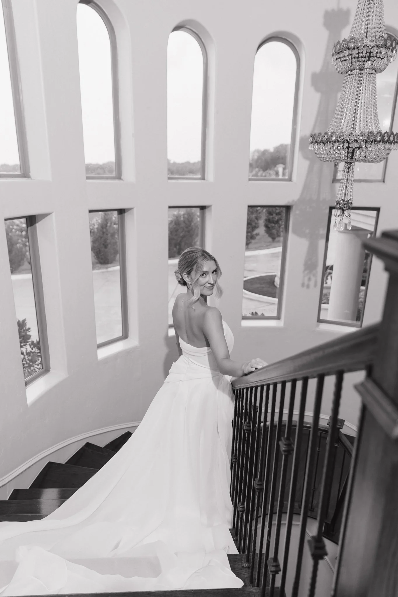 A woman in a wedding dress standing on a curved staircase, looking back over her shoulder, in a brightly lit room with large windows and a chandelier.