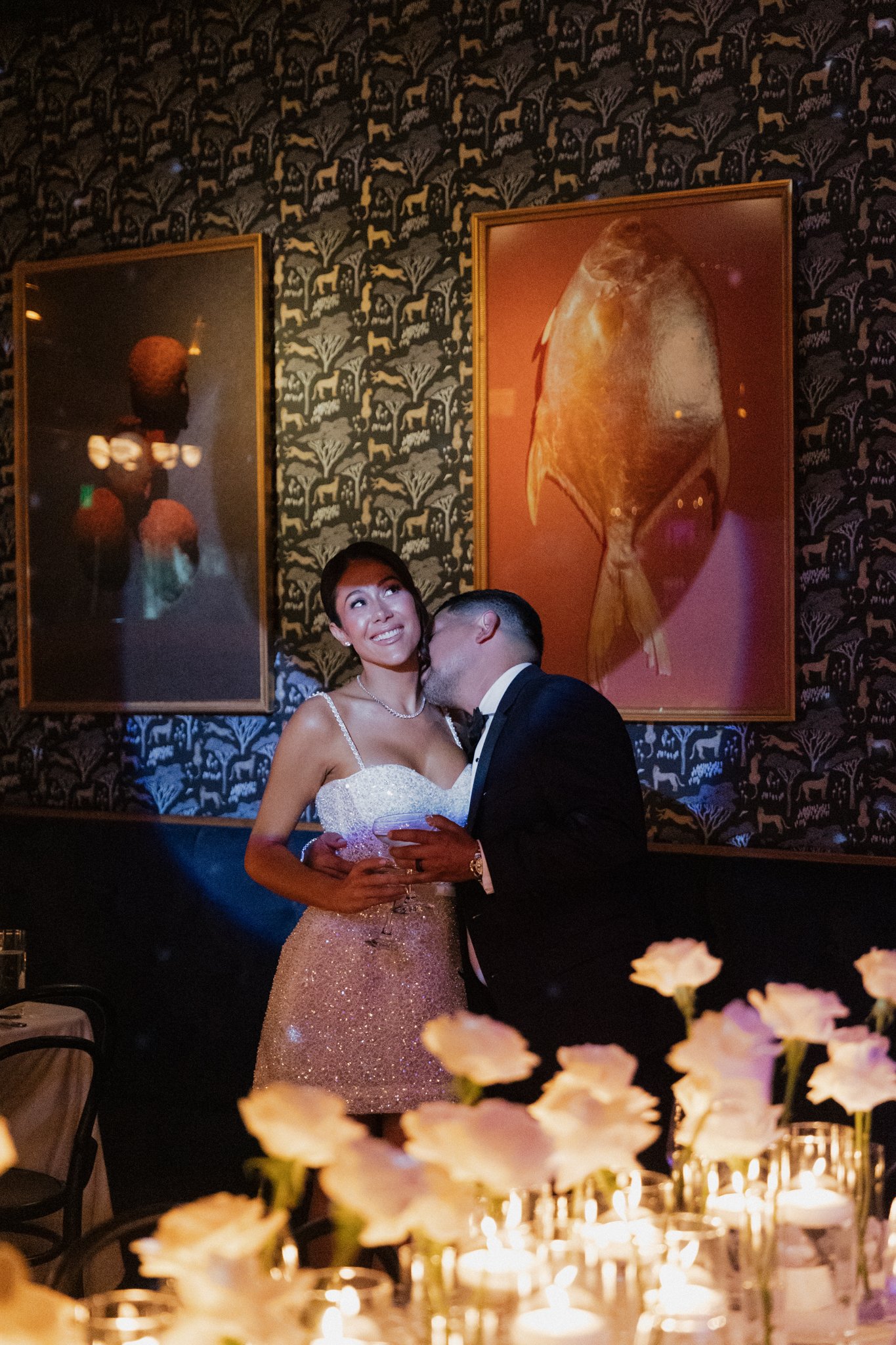 A newly married couple sharing a moment at their wedding reception, with candles and flowers in the foreground, and and modern art artwork on a patterned wall behind them.