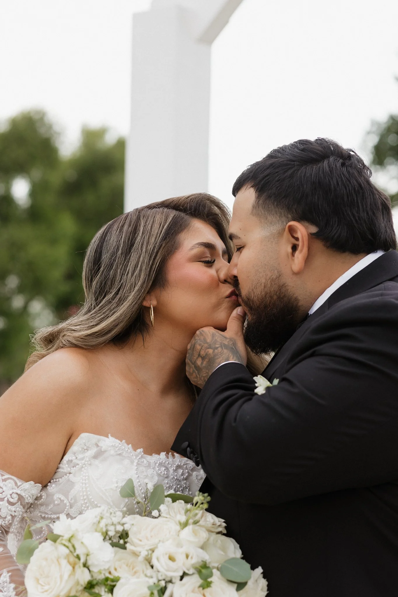 A bride and groom share a kiss outdoors, with the bride holding a bouquet of white flowers, during their wedding ceremony.