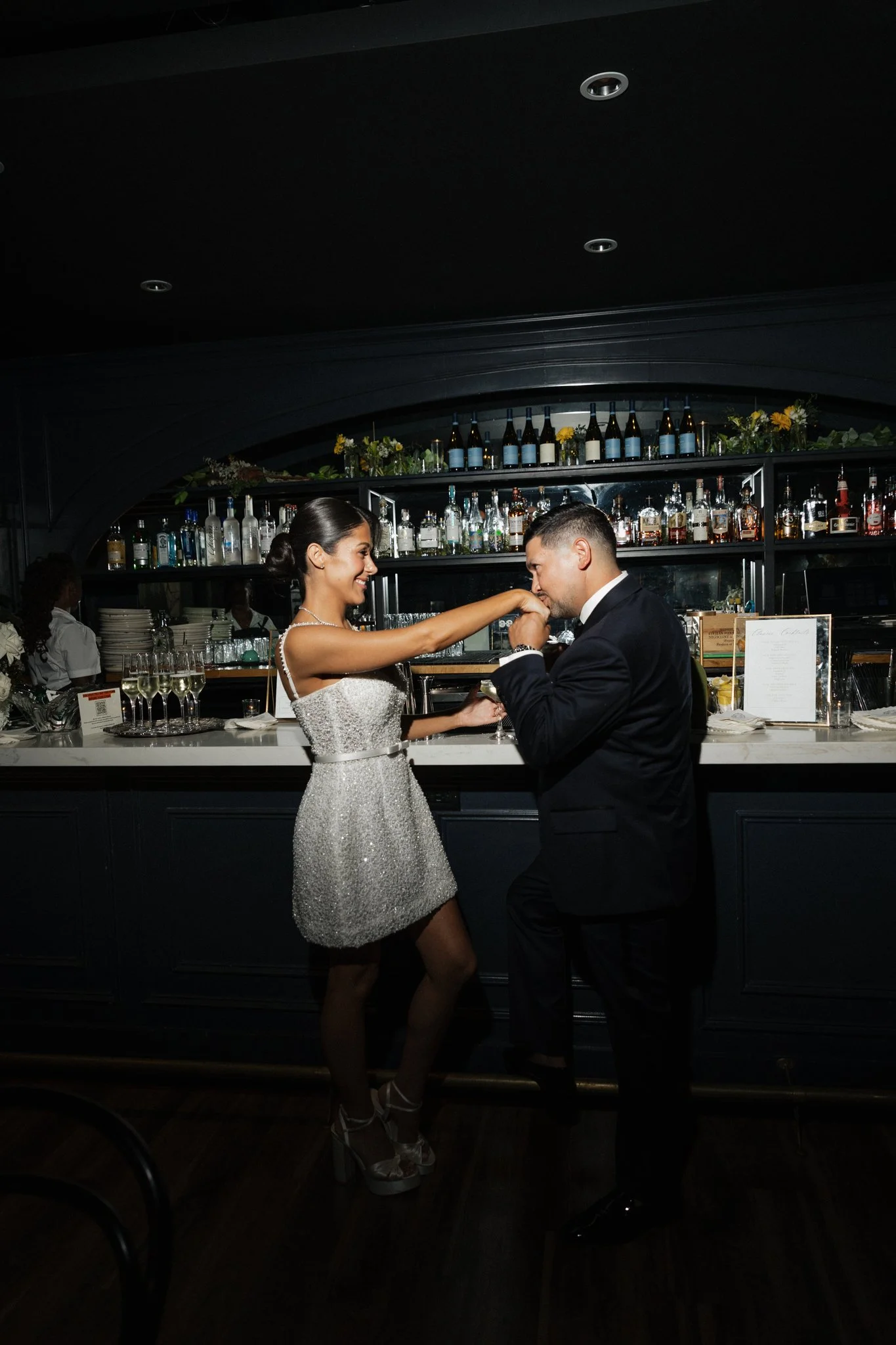 A couple dancing close together at a bar, with the woman wearing a sparkly dress and the man in a suit. The background shows a bar with bottles of alcohol and glasses.
