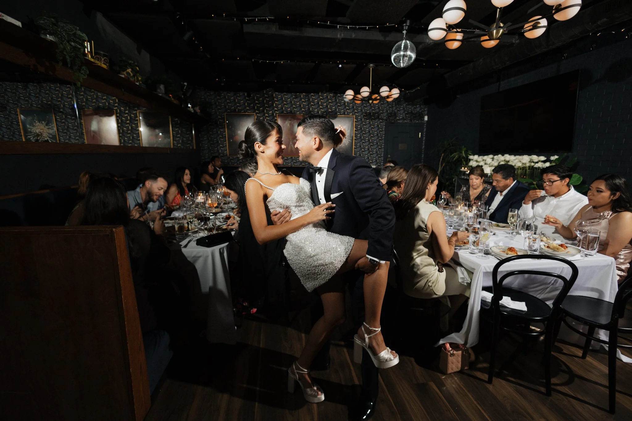 A bride and groom dancing at their wedding reception surrounded by seated guests, with a dark decorated interior and hanging lights.