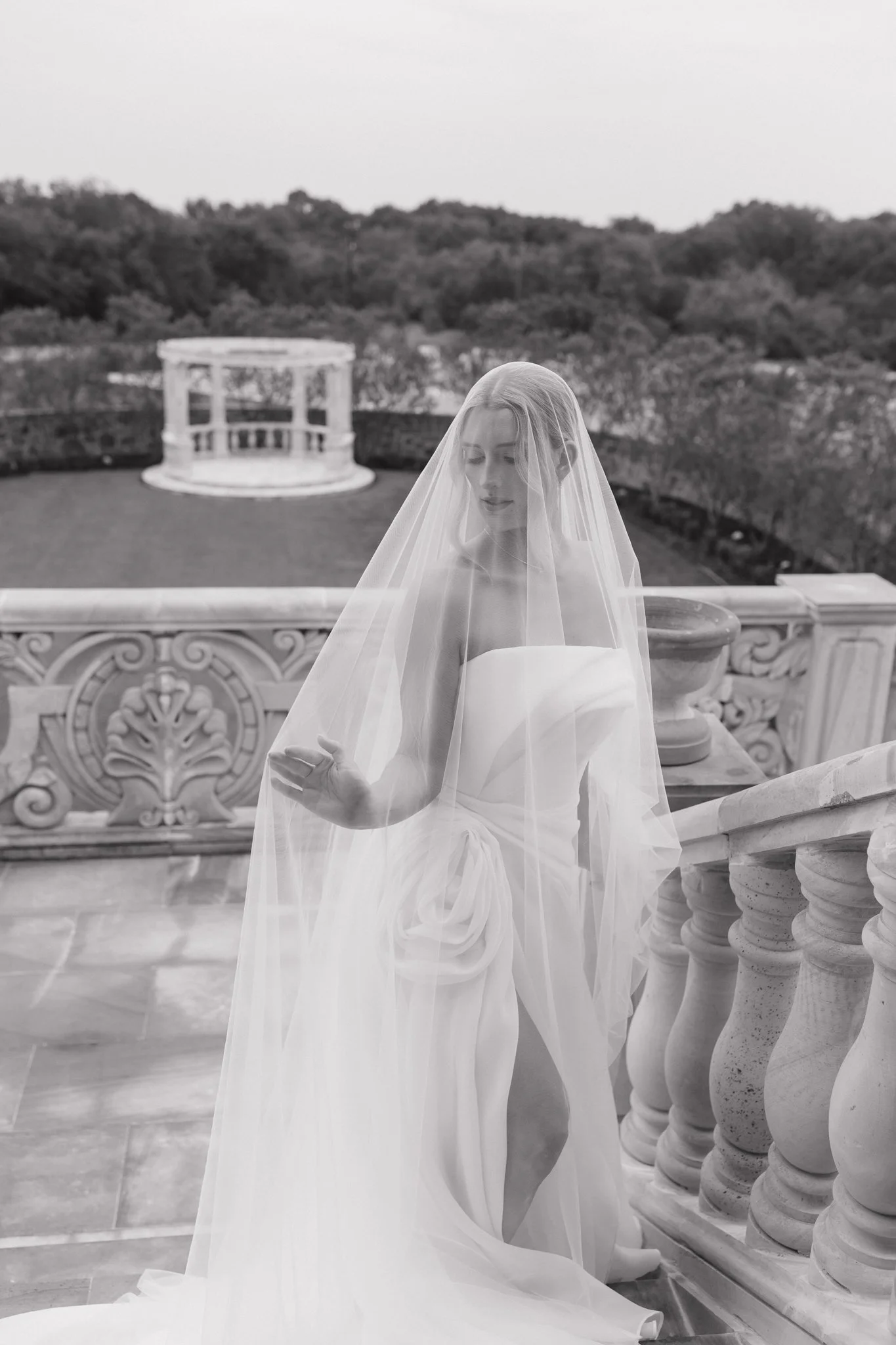 A bride in a strapless wedding gown and veil standing on a balcony with ornate balustrades, looking down and gently touching her dress.