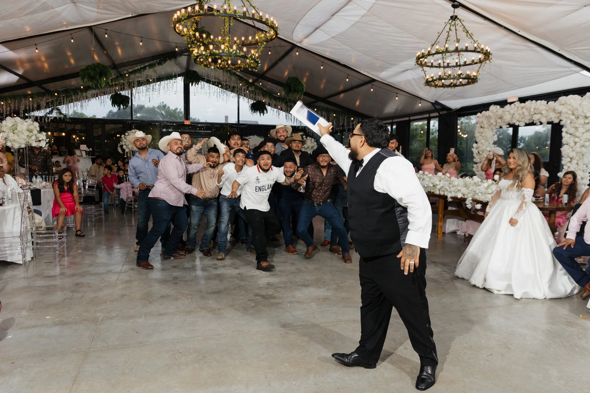 Man in black vest and white shirt taking selfie with group of guests, some wearing cowboy hats, at a wedding reception with decorated tent and floral arrangements.
