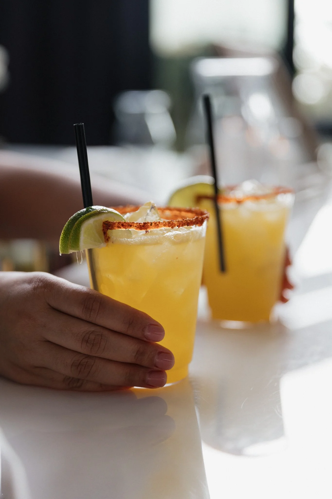 Two margarita cocktails with lime wedges and salted rims on a white table, one hand holding the front glass, blurred background.