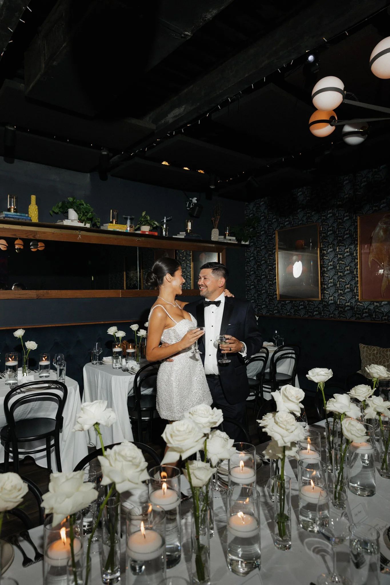A bride and groom celebrating at their wedding reception, standing close and smiling, with tables decorated with white roses and candles in the foreground, and modern black chairs and dark patterned walls in the background.