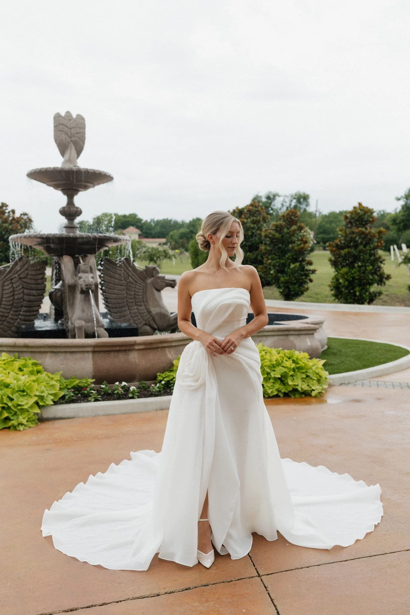 A bride in a strapless white wedding gown with a long train standing outdoors in front of a decorative fountain with cherub and angel sculptures.