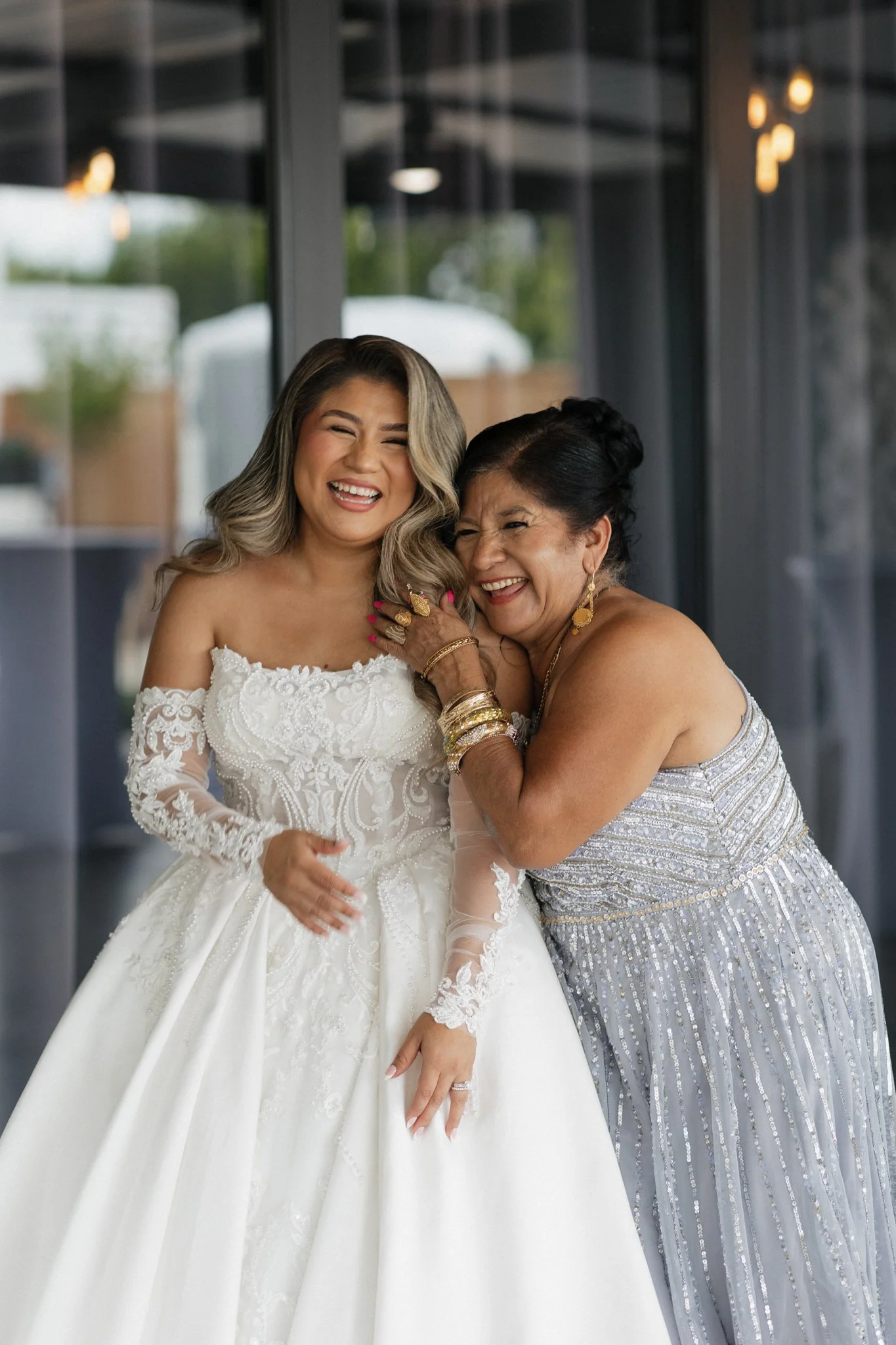 A bride in a white wedding dress laughing and smiling with an older woman in a silver dress, sharing a joyful moment.