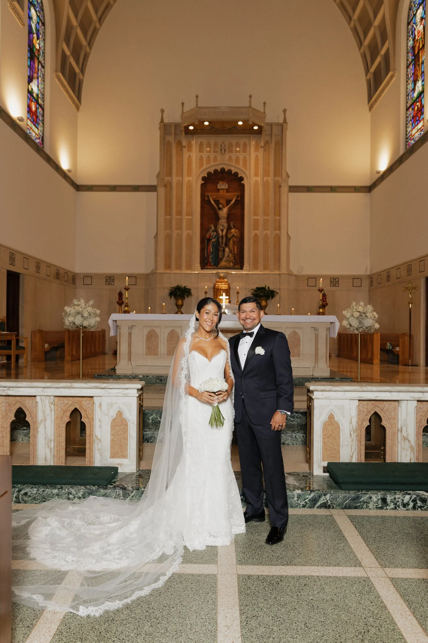 Bride and groom standing together inside a church altar area, smiling for a photo. The bride wears a white wedding gown with a long lace train and veil, holding a bouquet, and the groom wears a black tuxedo with a bow tie.