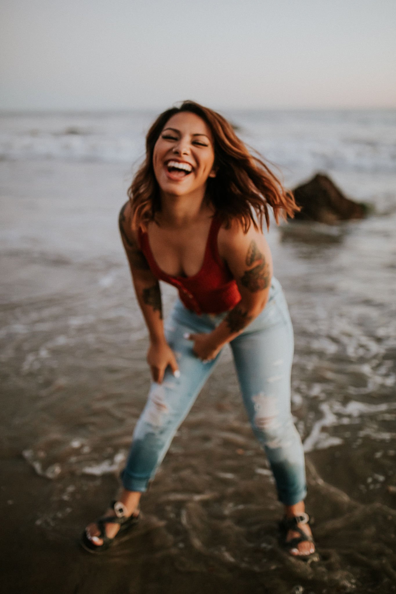 A woman with wavy hair, tattoos on her arms, wearing a red top, light blue distressed jeans, and sandals, smiling and laughing on a beach as the waves come up around her legs during sunset.