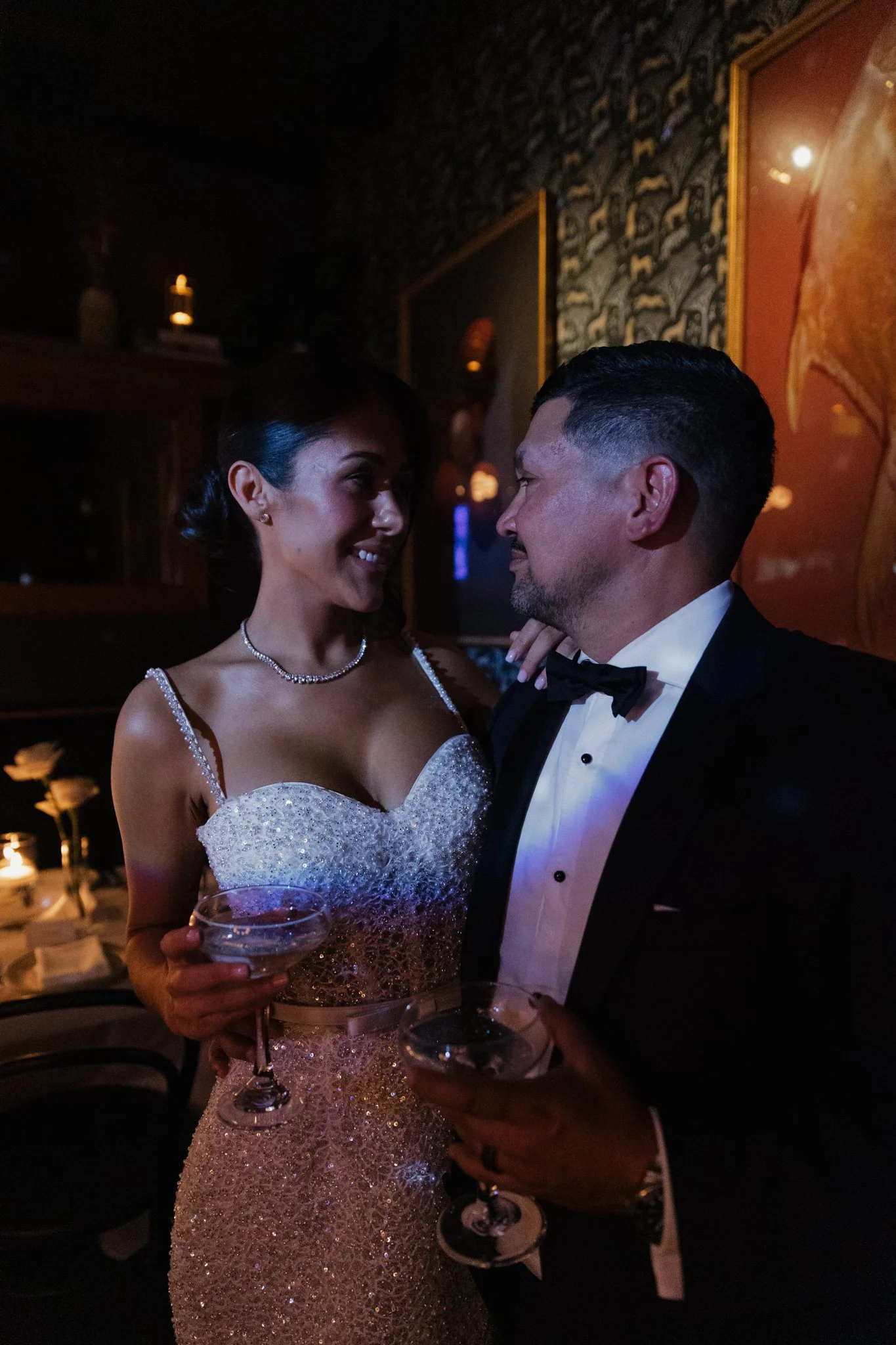 A bride and groom stand close together during their wedding reception, gazing into each other's eyes, the bride holding a glass of champagne, both dressed in formal wedding attire in a dimly lit room with elegant decor.