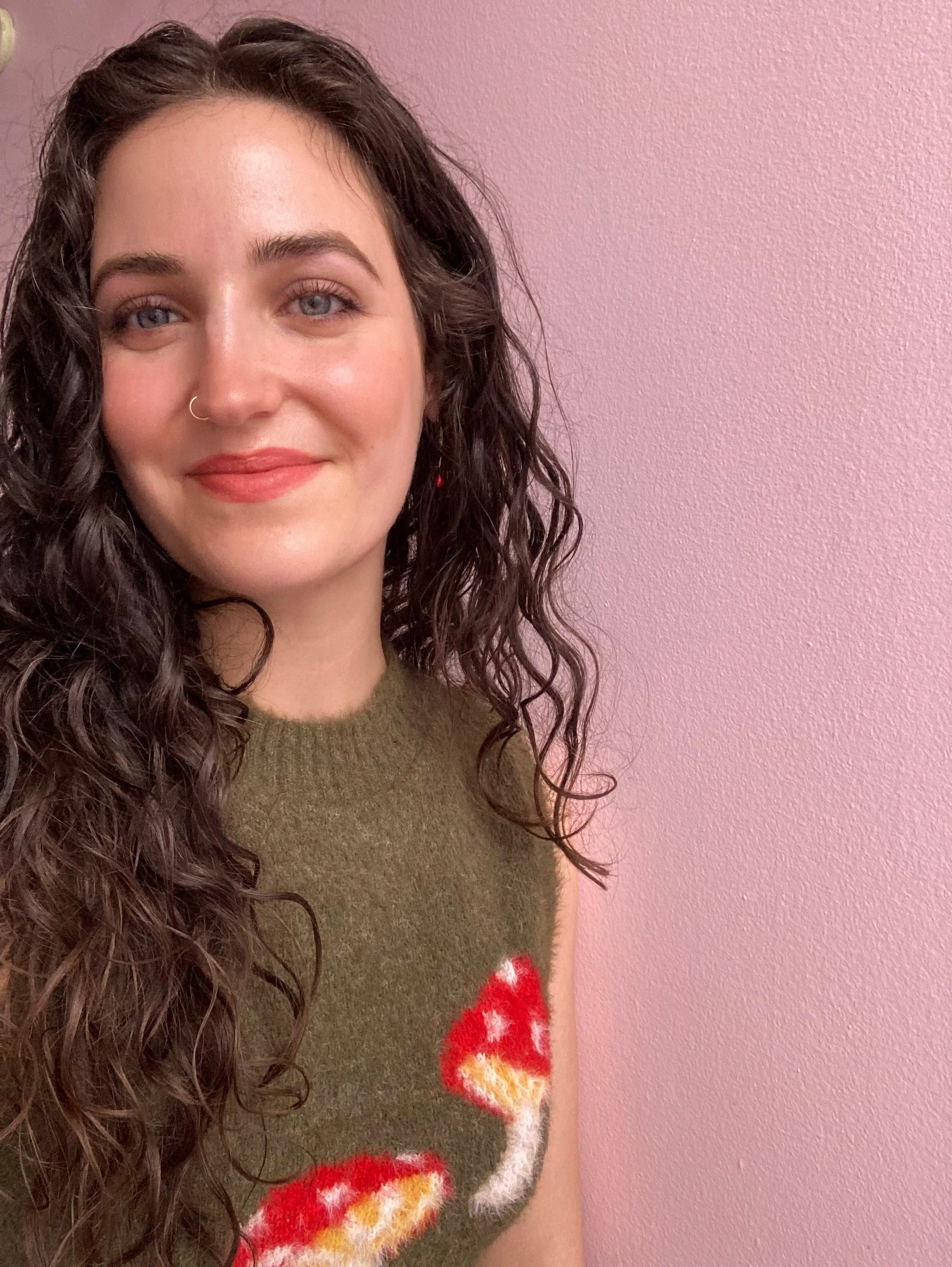 Close-up of a woman with dark curly hair, light skin, wearing a nose ring and a green sweater with a red and white pattern, standing against a pink wall.