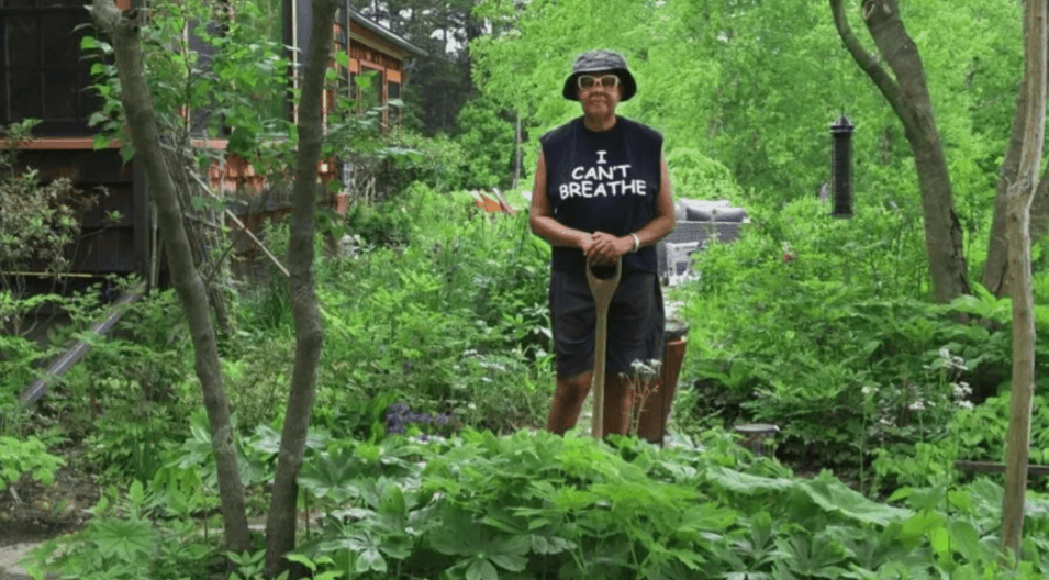 [Author Jamaica Kincaid stands outside in her garden, hands clasped in front of a shovel. She wears a black cut-off t-shirt with the phrase, “I Can’t Breathe” printed on the front.]