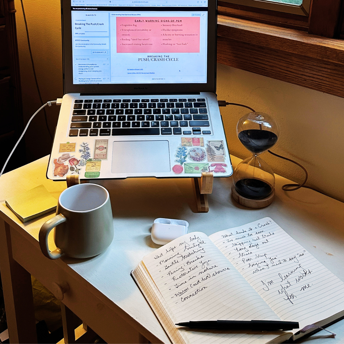 desk with computer, journal, coffee cup, AirPods, sandtimer