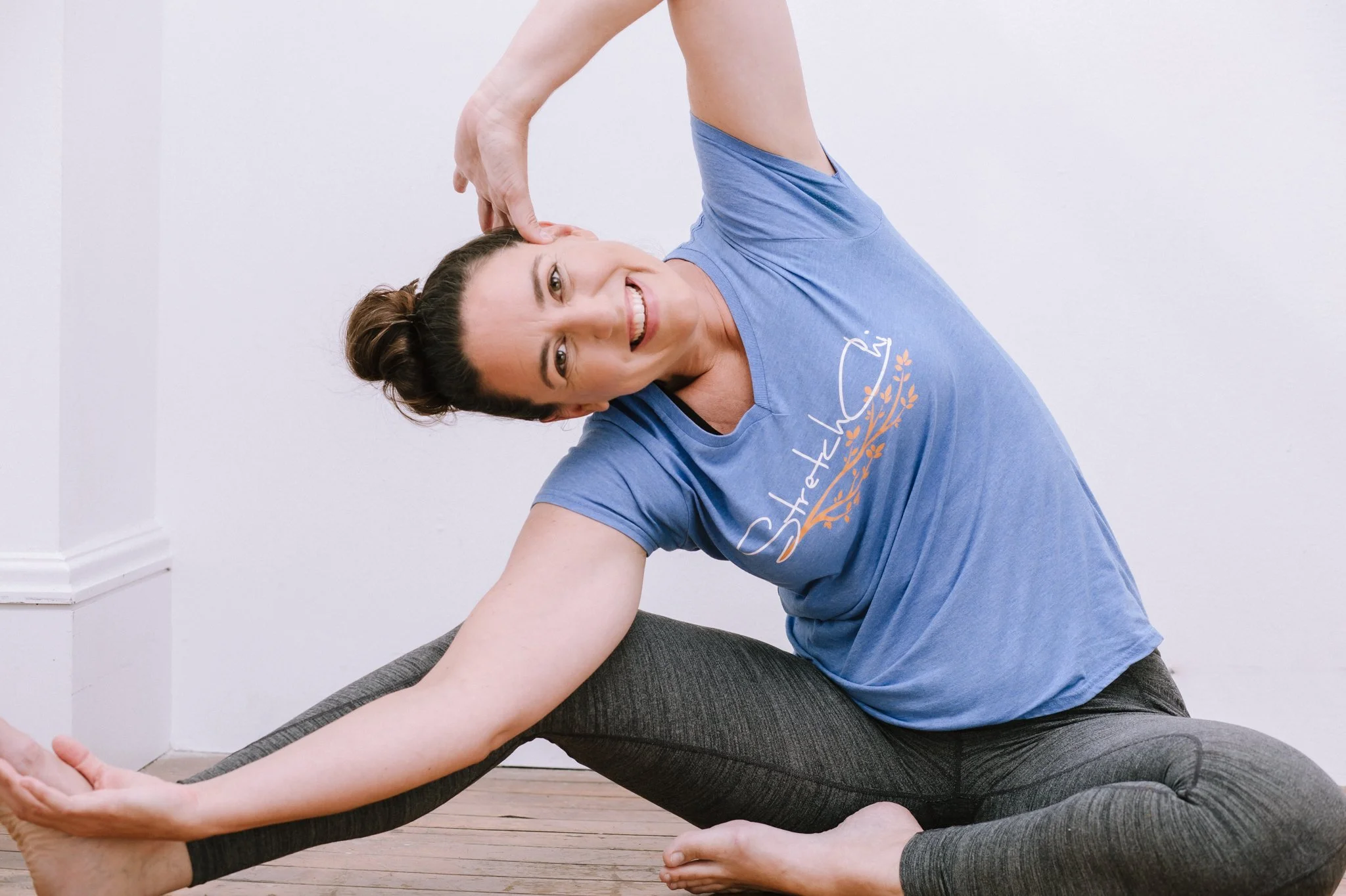 Carrie Collins, owner of Stretch Chi,  practicing resistance stretching in a seated side stretch pose, smiling, wearing a blue t-shirt and gray leggings, in a room with white walls and wooden floor.
