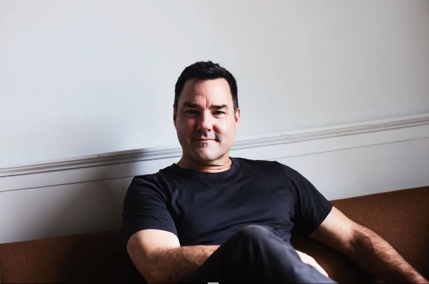 Man sitting on a sofa in a relaxed pose, wearing a black shirt, with a plain wall in the background.