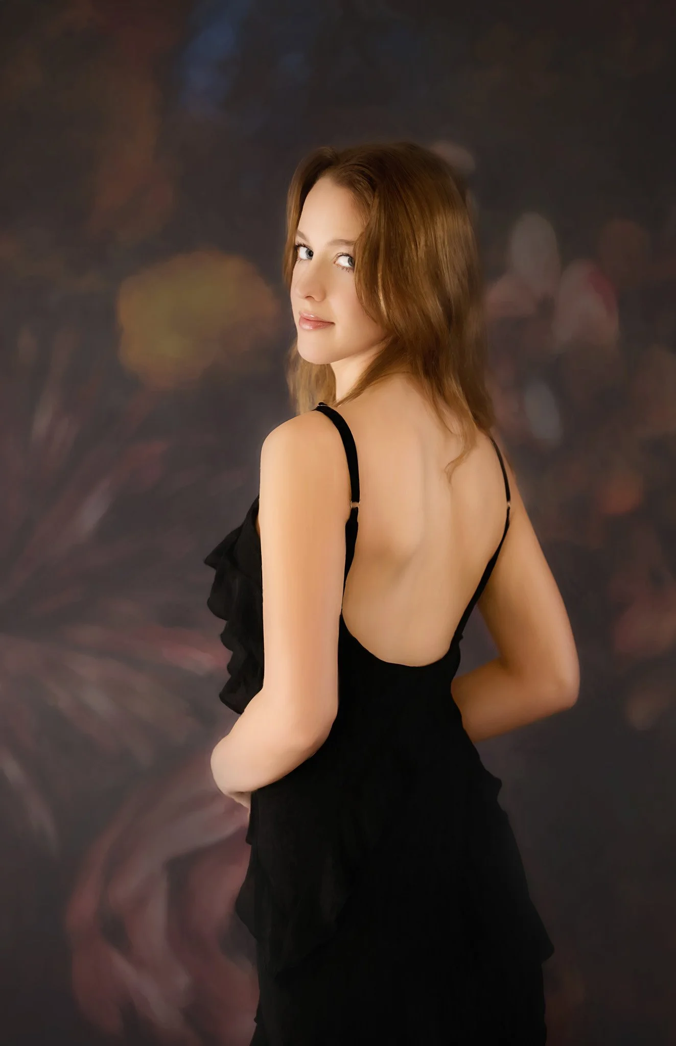 A young woman with long red hair wearing a black backless dress, standing against a dark, softly blurred background.