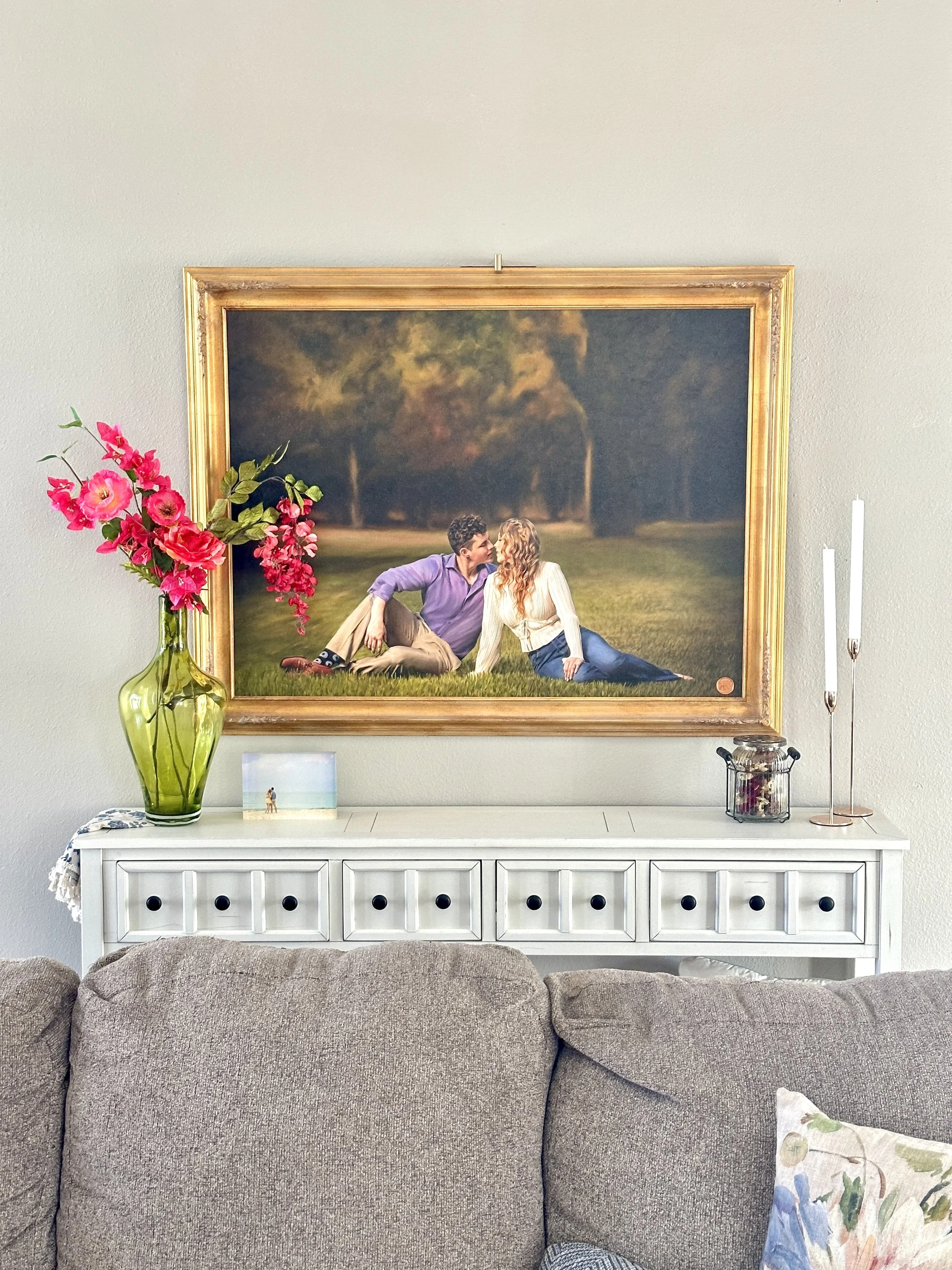 Living room with a gray upholstered couch in front of a white sideboard. On the sideboard, there is a large green vase with pink flowers, a small framed picture of a beach, and two white candles on rose gold candleholders. A large framed painting of 
