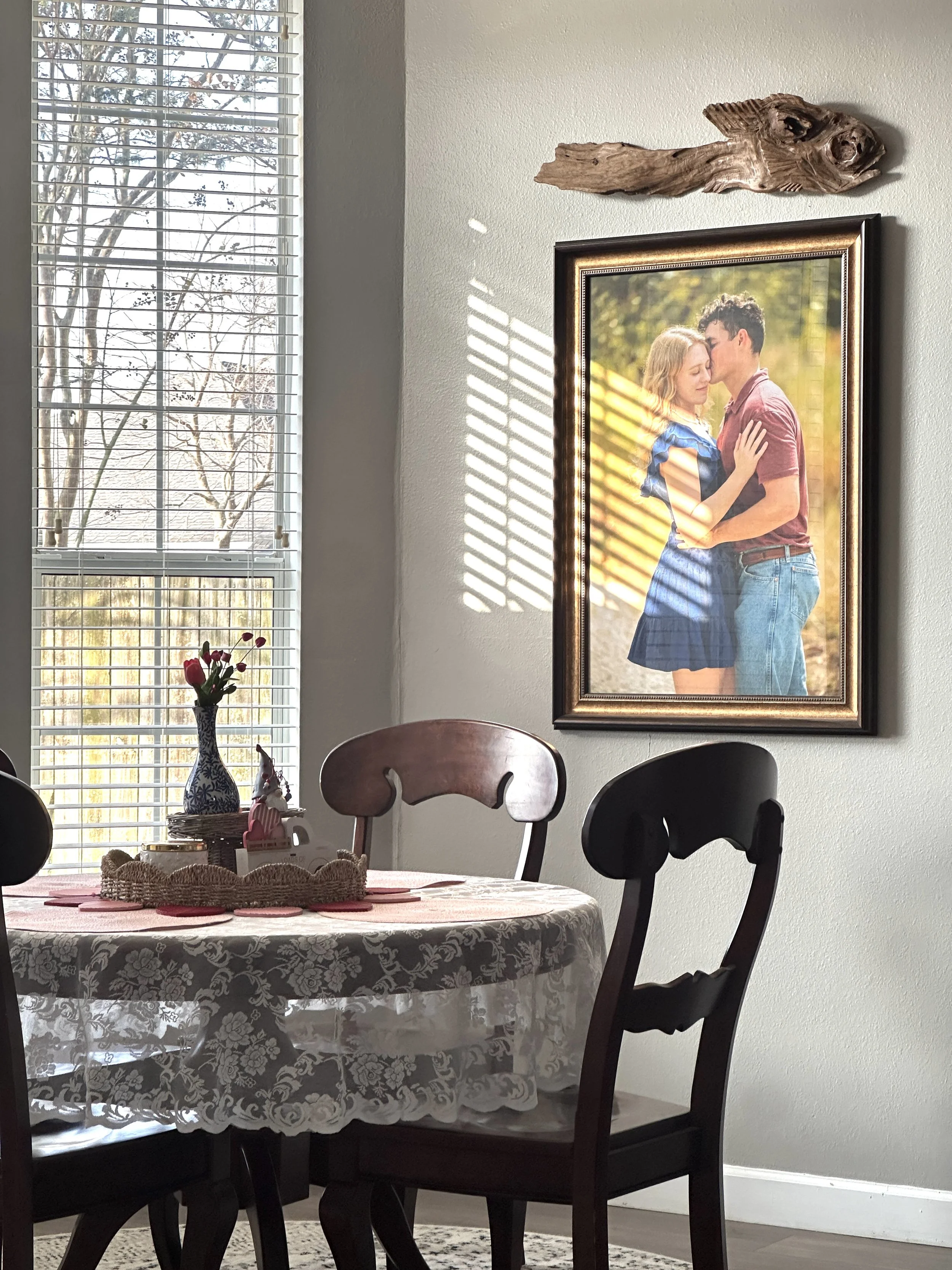 A dining room corner with a table covered by a lace tablecloth, a vase with tulips, and a framed photograph of a couple embracing on the wall, with a wooden decorative piece above it. Sunlight streams through a window with blinds.