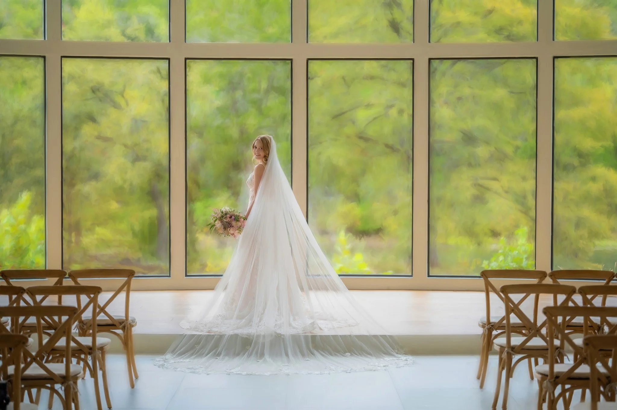 A bride in a white wedding gown holding a bouquet of flowers, posing indoors near large windows showing green trees outside.